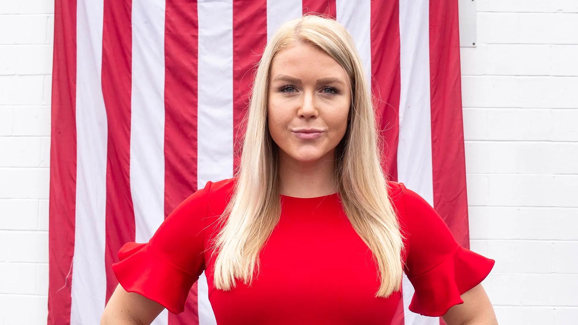 O blondă cu părul lung, într-o rochie roșie, stă pe fundalul steagului american