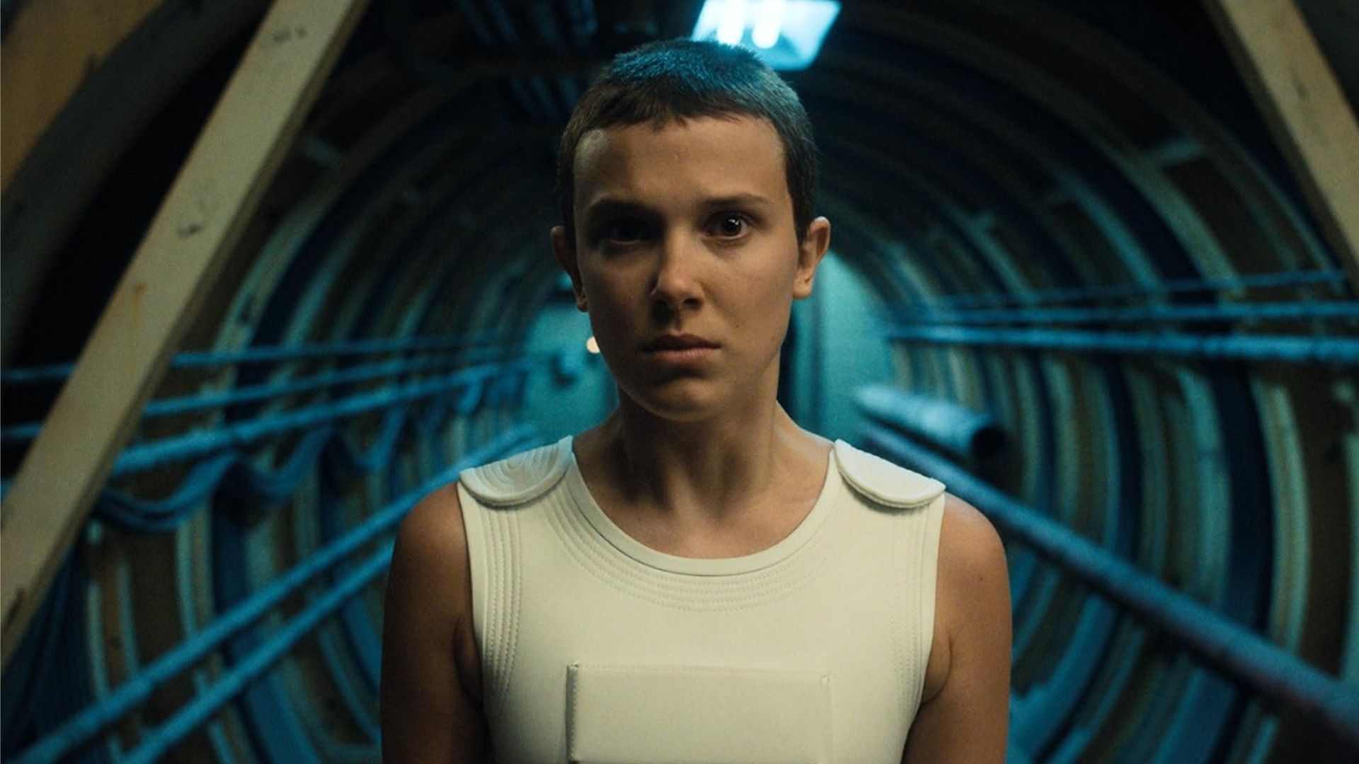 Millie Bobby Brown cu o tunsoare scurtă stă într-un tunel și privește către cameră