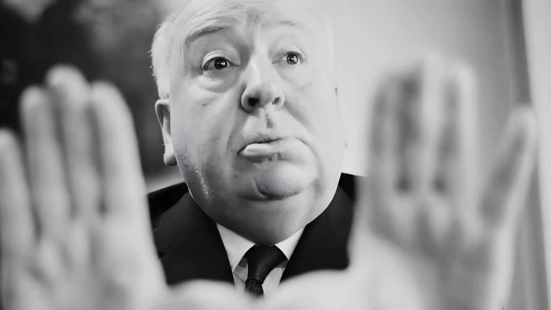 Un prim-plan al feței lui Alfred Hitchcock, cu mâinile întinse în semn de oprire