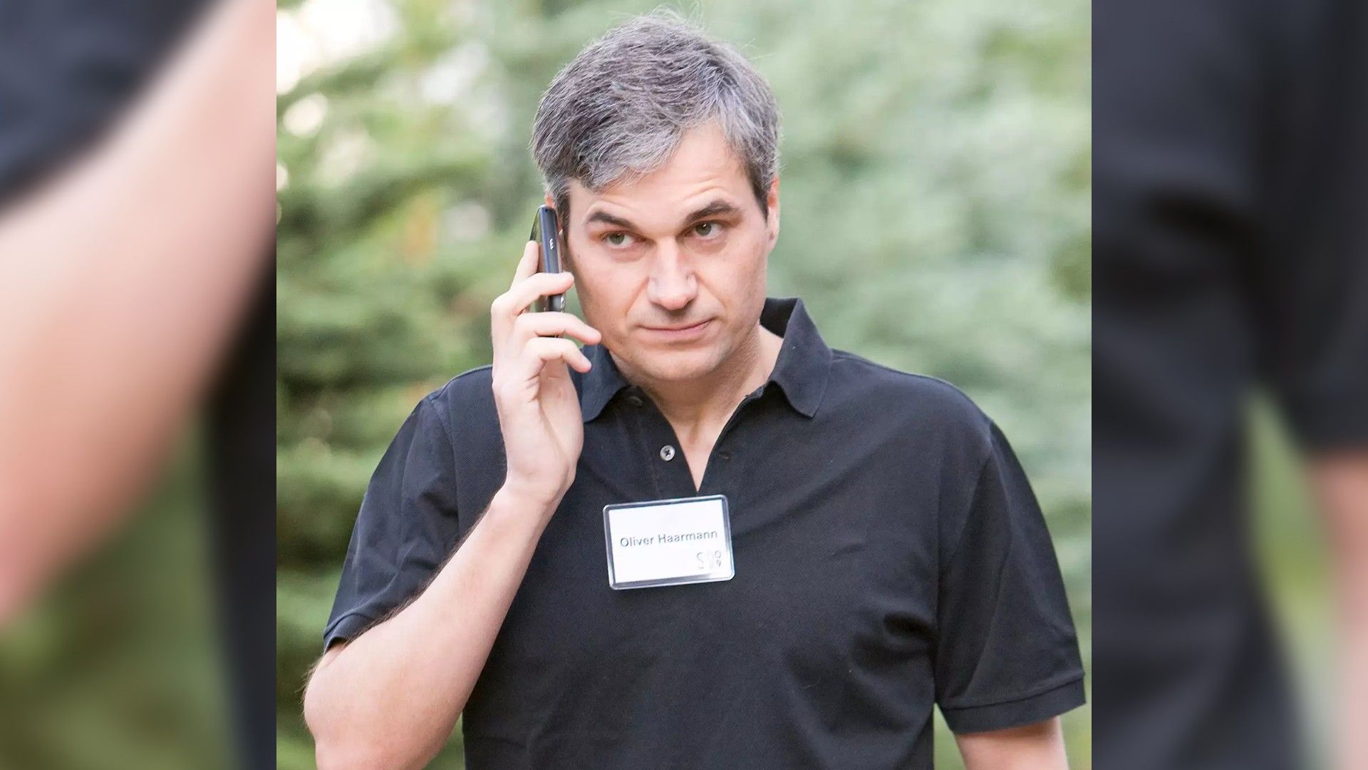 Un bărbat în cămașă neagră cu insignă vorbind la telefon pe fondul unor copaci verzi, arătând concentrat