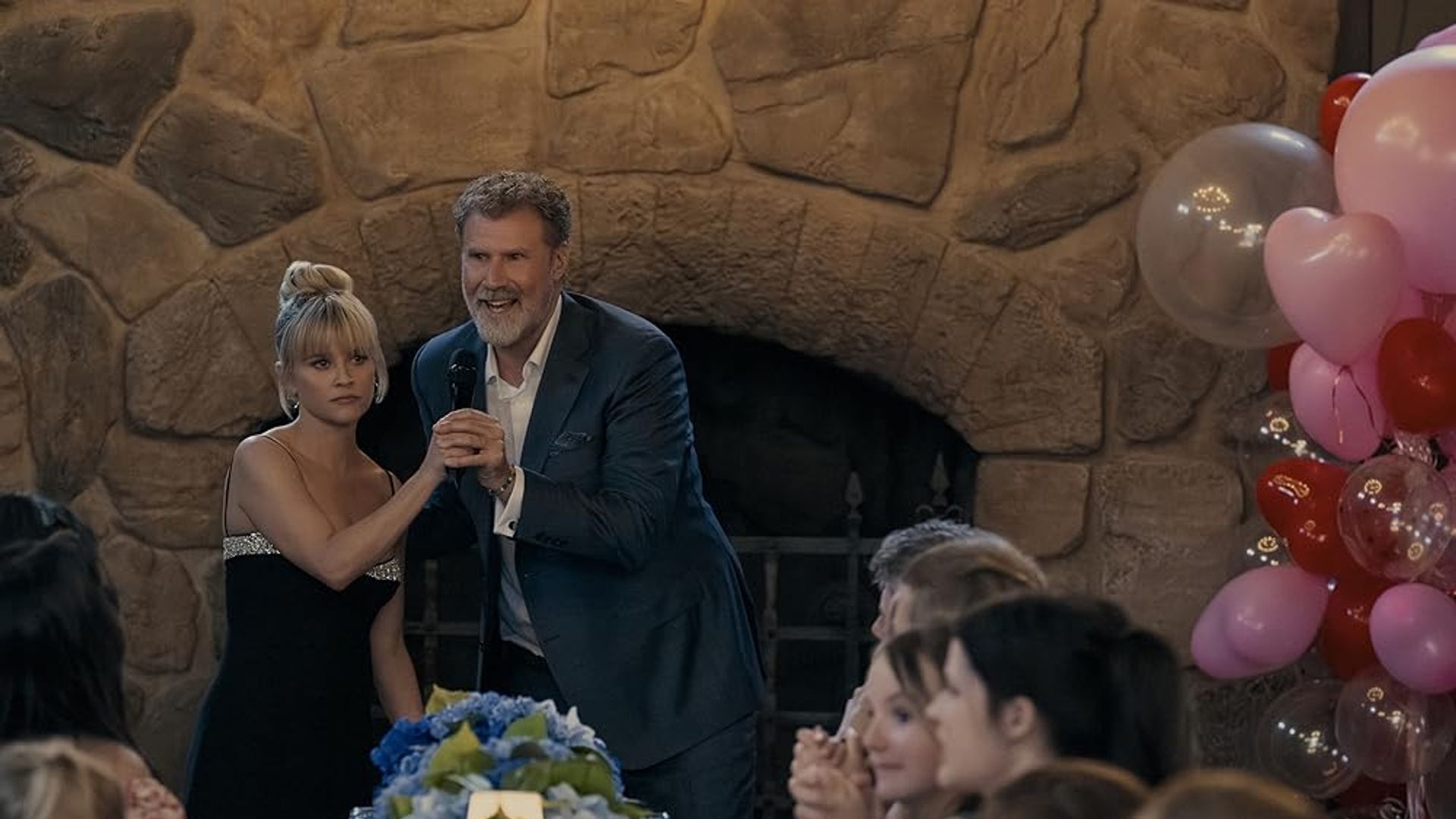 Will Ferrell în costum cu microfon ține de mână pe Reese Witherspoon în rochie neagră pe fondul unei șeminee, unei mese festive și baloanelor