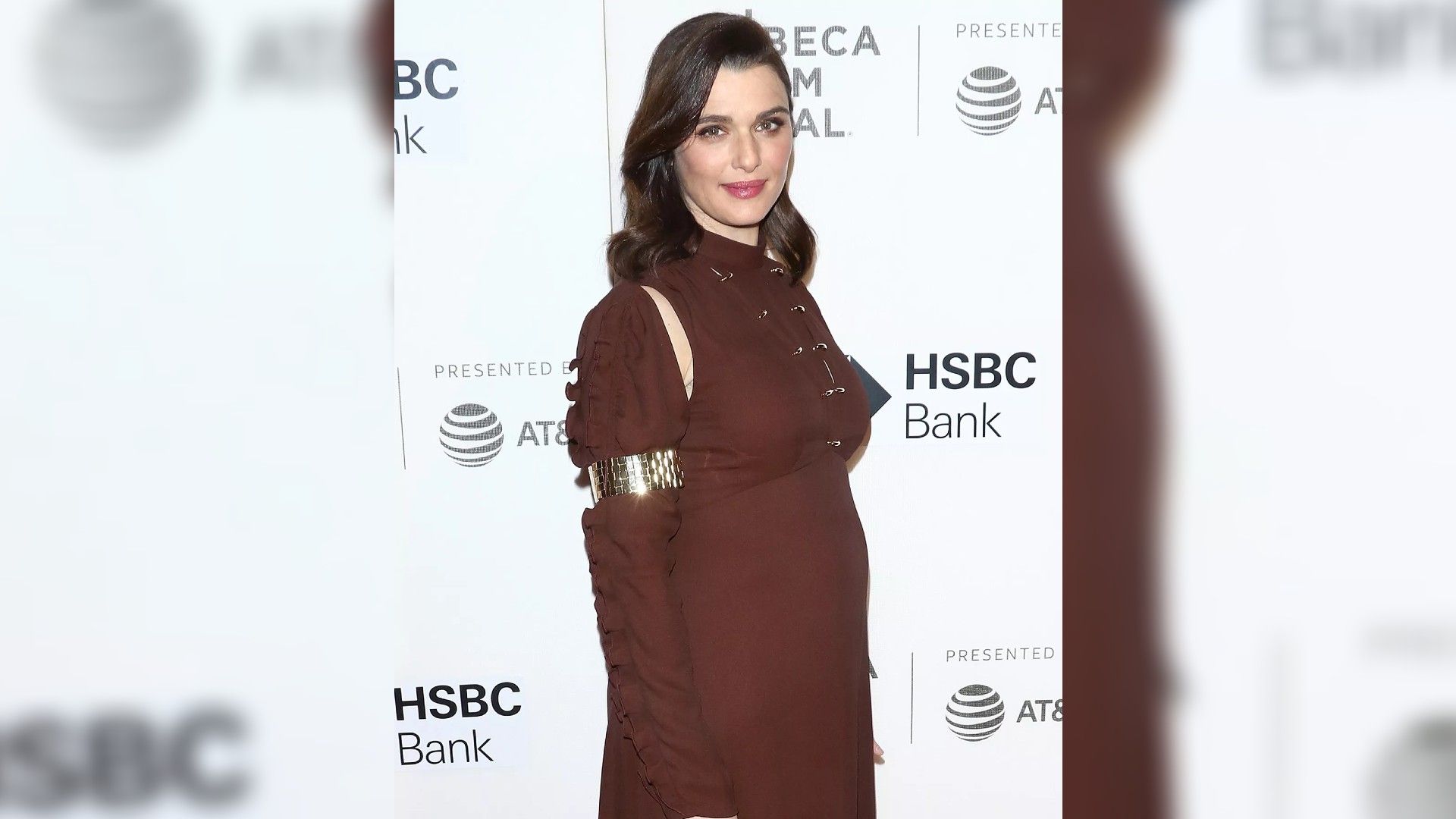 Rachel Weisz într-o rochie burgundy, în picioare, arătând clar că așteaptă un copil