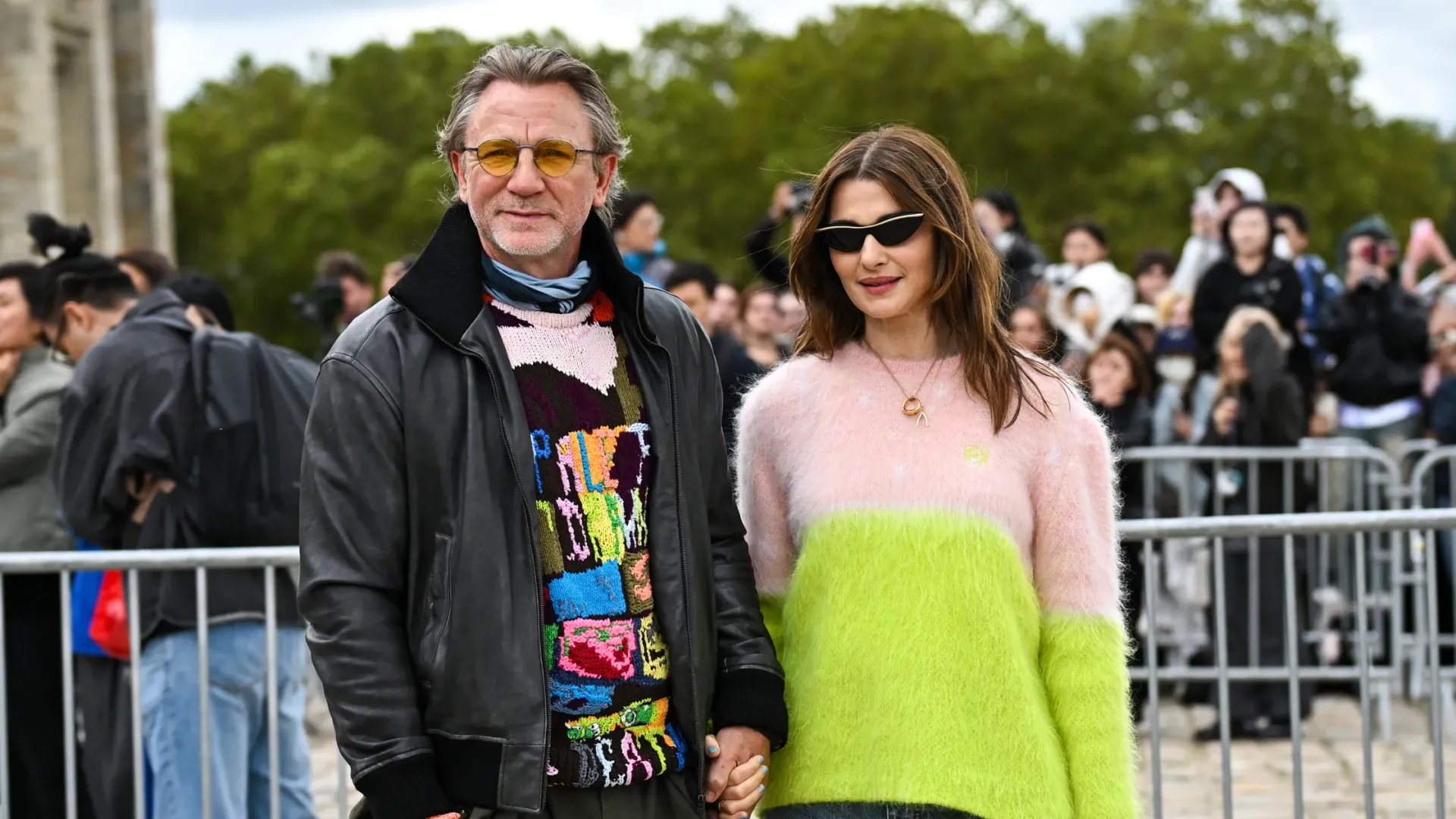 Rachel Weisz într-un pulover alb și verde și Daniel Craig într-o jachetă de piele neagră și un pulover colorat, ținându-se de mână