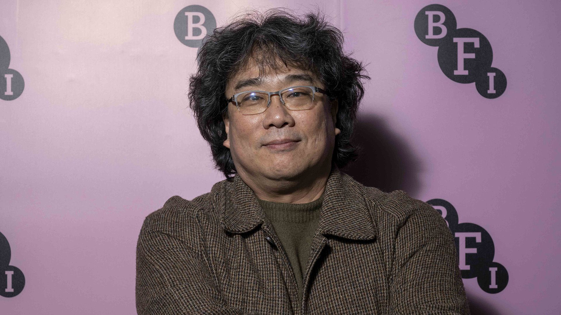 Directorul Bong Joon Ho într-o jachetă maro și ochelari pe un fundal liliachiu