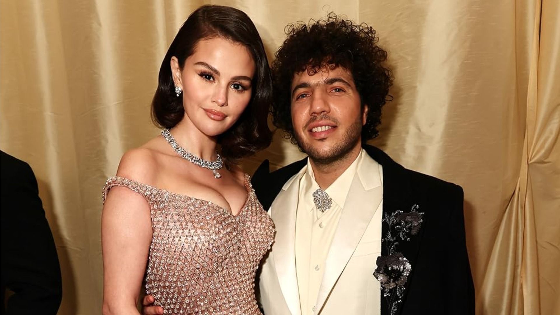 Actrița Selena Gomez și logodnicul ei, producătorul muzical Benny Blanco, se îmbrățișează și zâmbesc la cameră pe un fundal auriu