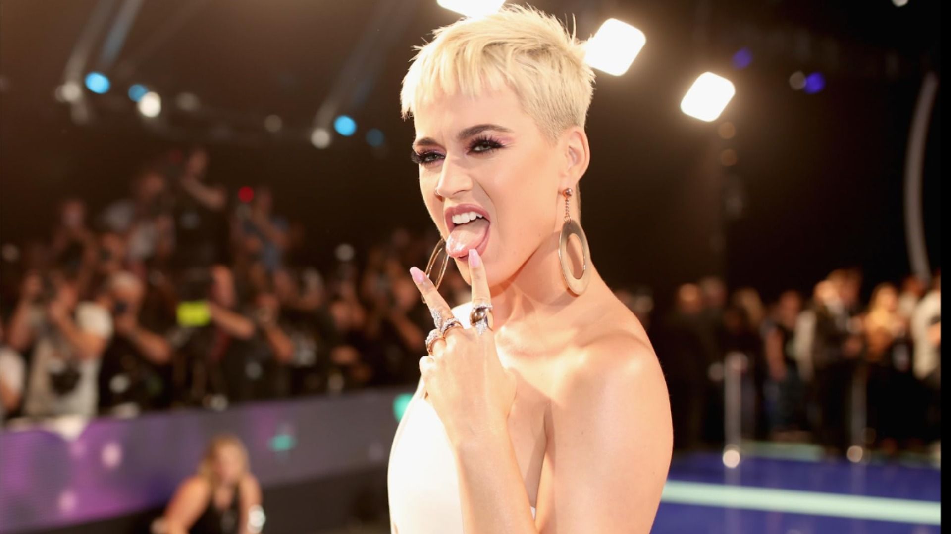 Cântăreața Katy Perry scoate limba și ridică două degete într-o rochie albă pe fundalul publicului