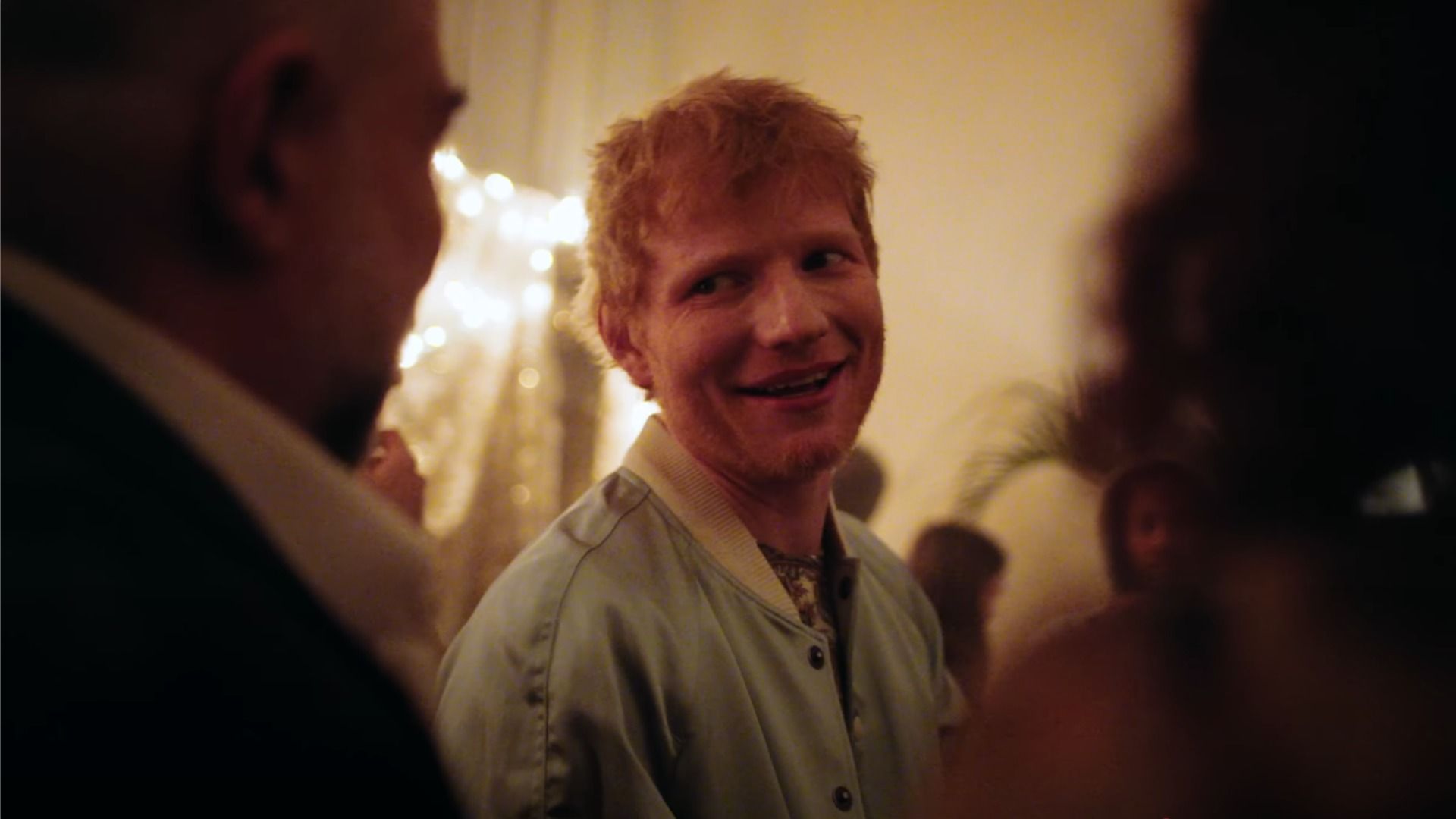 Cântărețul Ed Sheeran privește și zâmbește în lumina galben-portocalie, lângă siluete de oameni