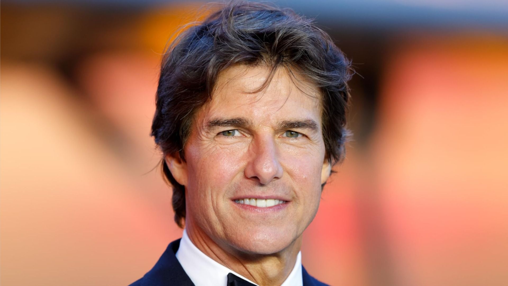 Actorul Tom Cruise pozează într-un costum clasic și zâmbește la cameră pe un fundal blurat