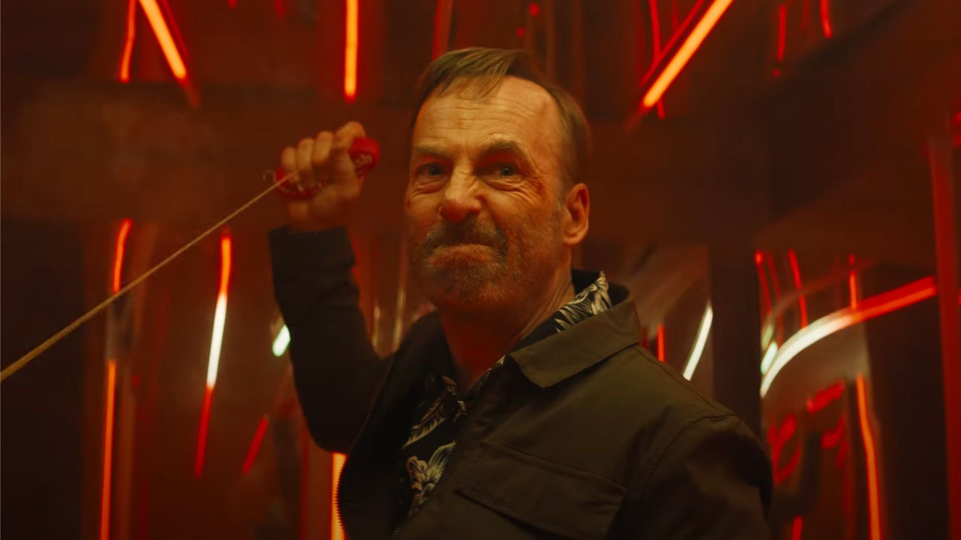 Actorul Bob Odenkirk într-o jachetă neagră ține o funie în mâini și privește furios în direcția luminii roșii