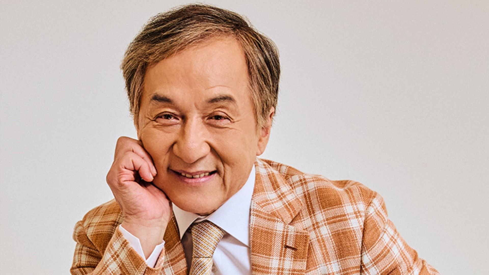 Actorul Jackie Chan într-o jachetă maro în carouri, stă sprijinit pe mână și zâmbește la cameră pe un fundal alb