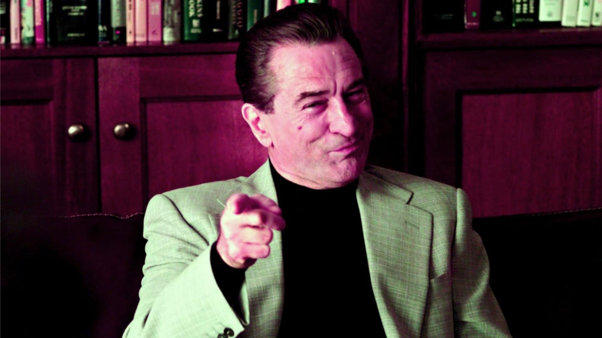 Actorul Robert De Niro într-o jachetă gri arătând cu degetul arătător și zâmbind pe fundalul unei biblioteci