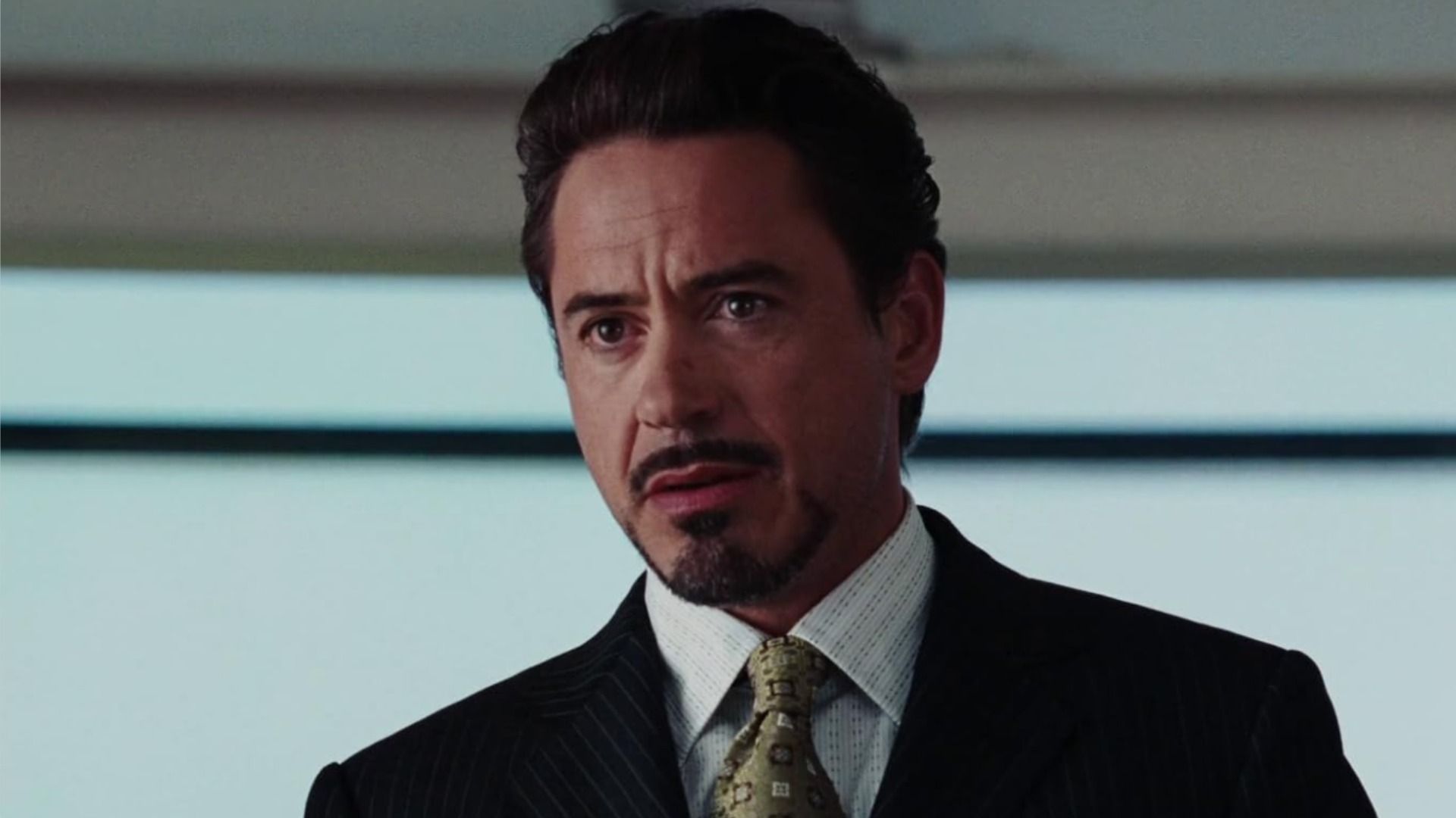 Actorul Robert Downey Jr. cu mustață și barbă stă într-un costum clasic și privește în lateral pe fundalul unei ferestre