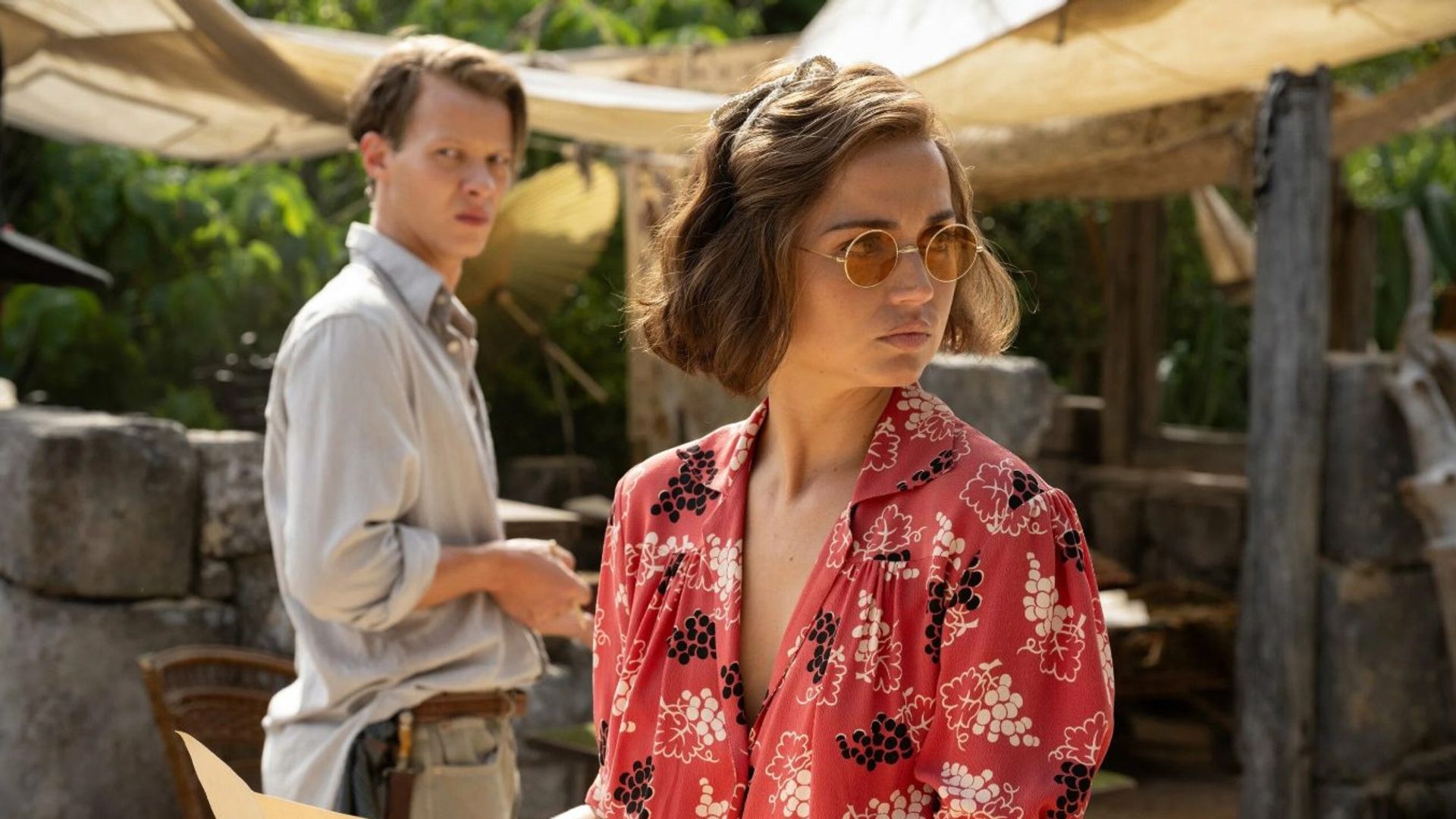 Ana de Armas într-o bluză roșie cu imprimeu și ochelari de soare ține o hartă, stând lângă un bărbat într-o cămașă deschisă la culoare, scena fiind amplasată în fața unor ruine arheologice