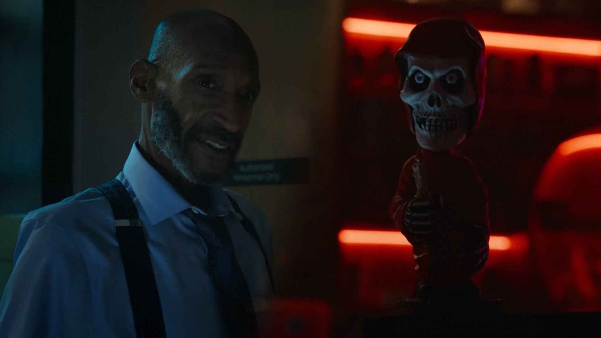 Actorul Tony Todd într-o cămașă albă cu bretele, stând în unghi și privind lateral pe fundalul unui birou