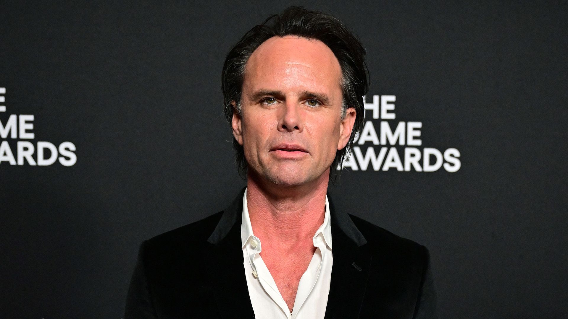 Actorul Walton Goggins într-o jachetă neagră și cămașă albă cu gulerul descheiat pe un fundal negru