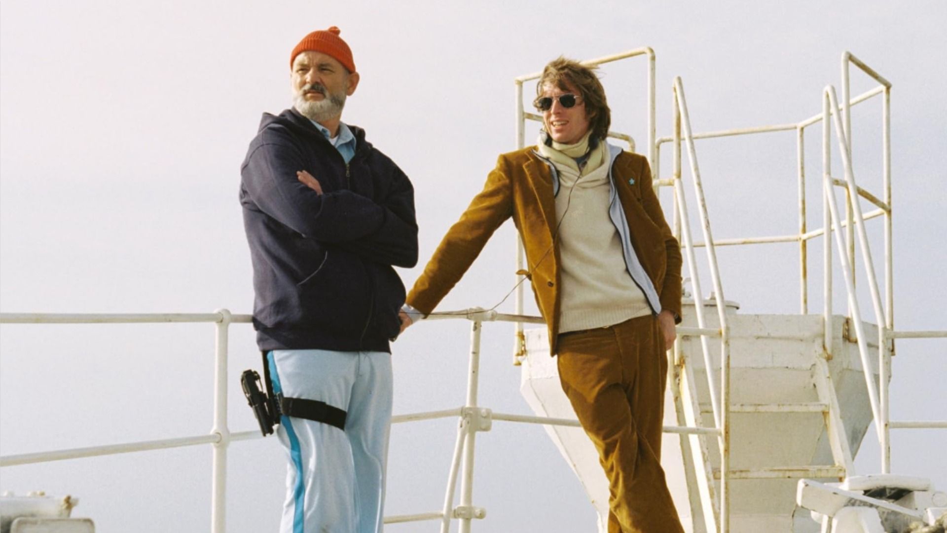 Actorul Bill Murray într-o pălărie roșie și regizorul Wes Anderson într-o jachetă maro, ținându-se de balustrada navei pe fundalul cerului