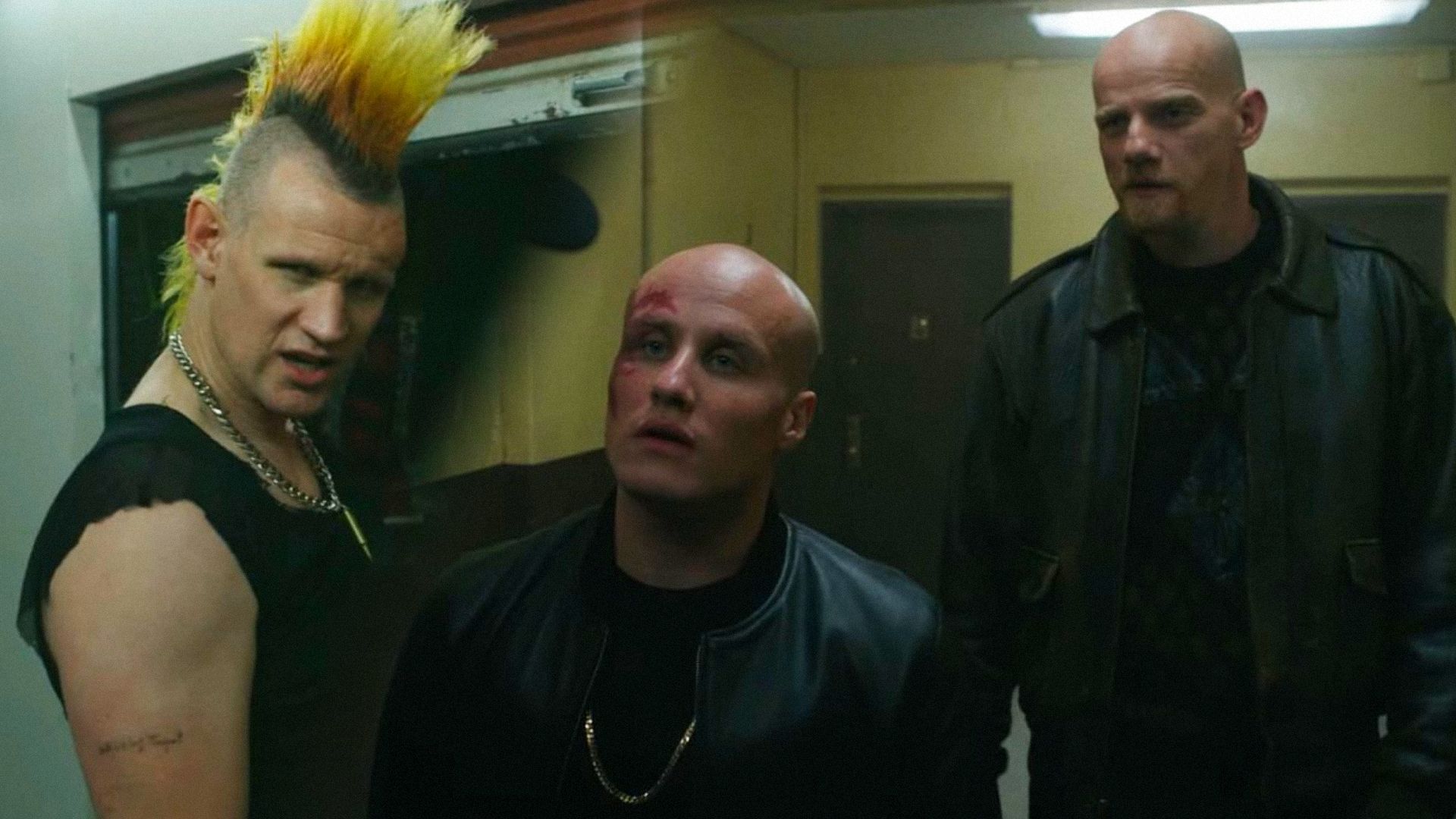 Actorul Matt Smith cu un mohawk privește în lateral, actorii Nikita Kukushkin și Yuri Kolokolnikov în ținute de piele stau pe hol și privesc în lateral