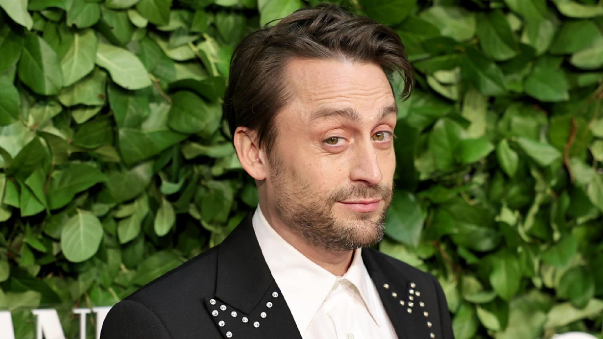Kieran Culkin looking ironically at the camera against a wall of leaves
