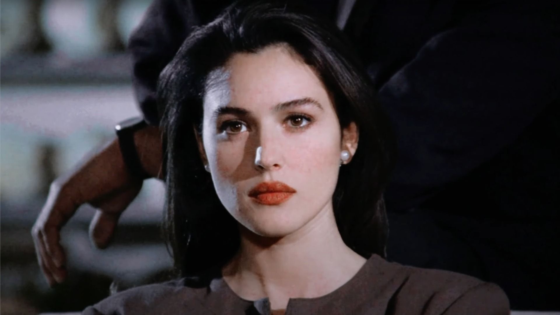 Actrița Monica Bellucci cu ruj roșu pe buze privește în lateral pe fundalul unui bărbat în costum
