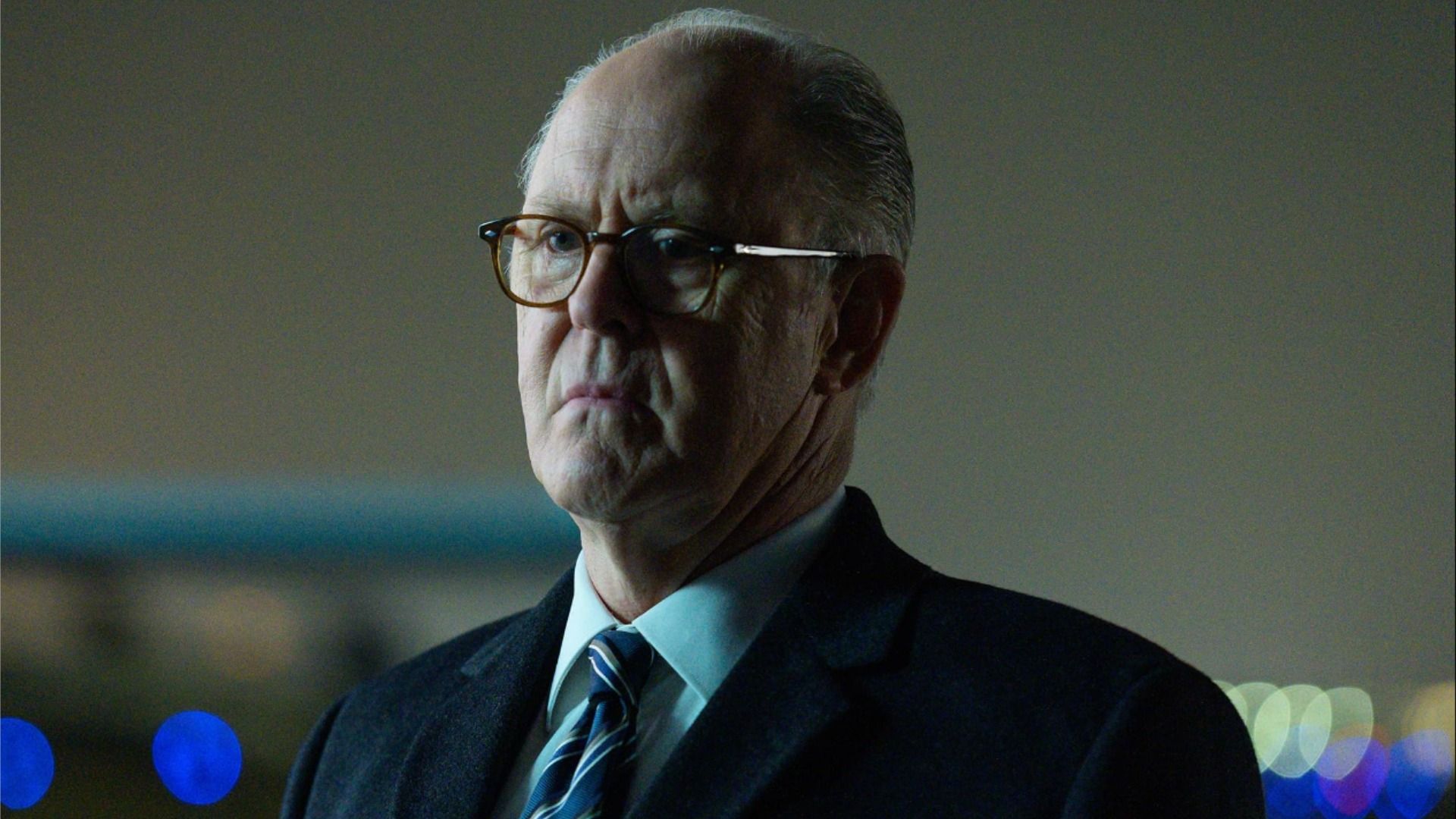 Actorul John Lithgow în costum și ochelari stă impasibil și privește în lateral