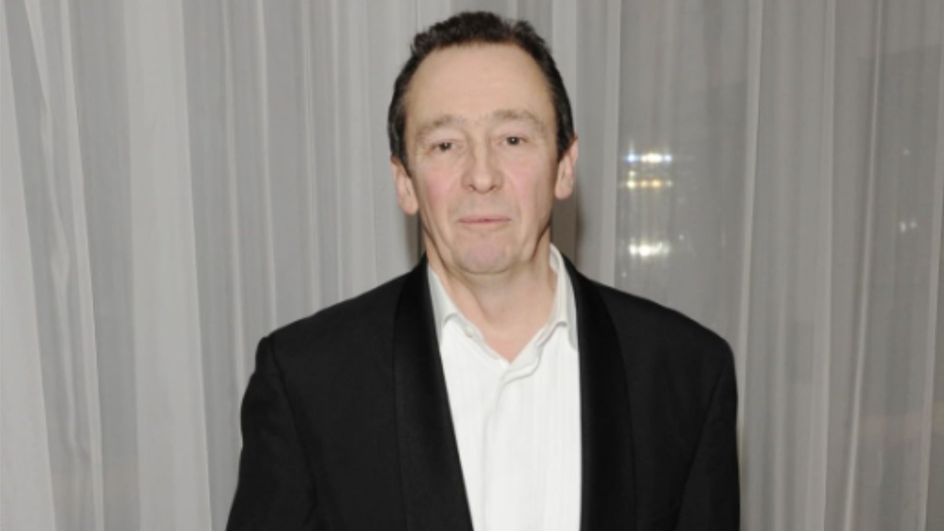 Actorul Paul Whitehouse stă într-un costum negru pe un fundal de tulle alb și privește la cameră