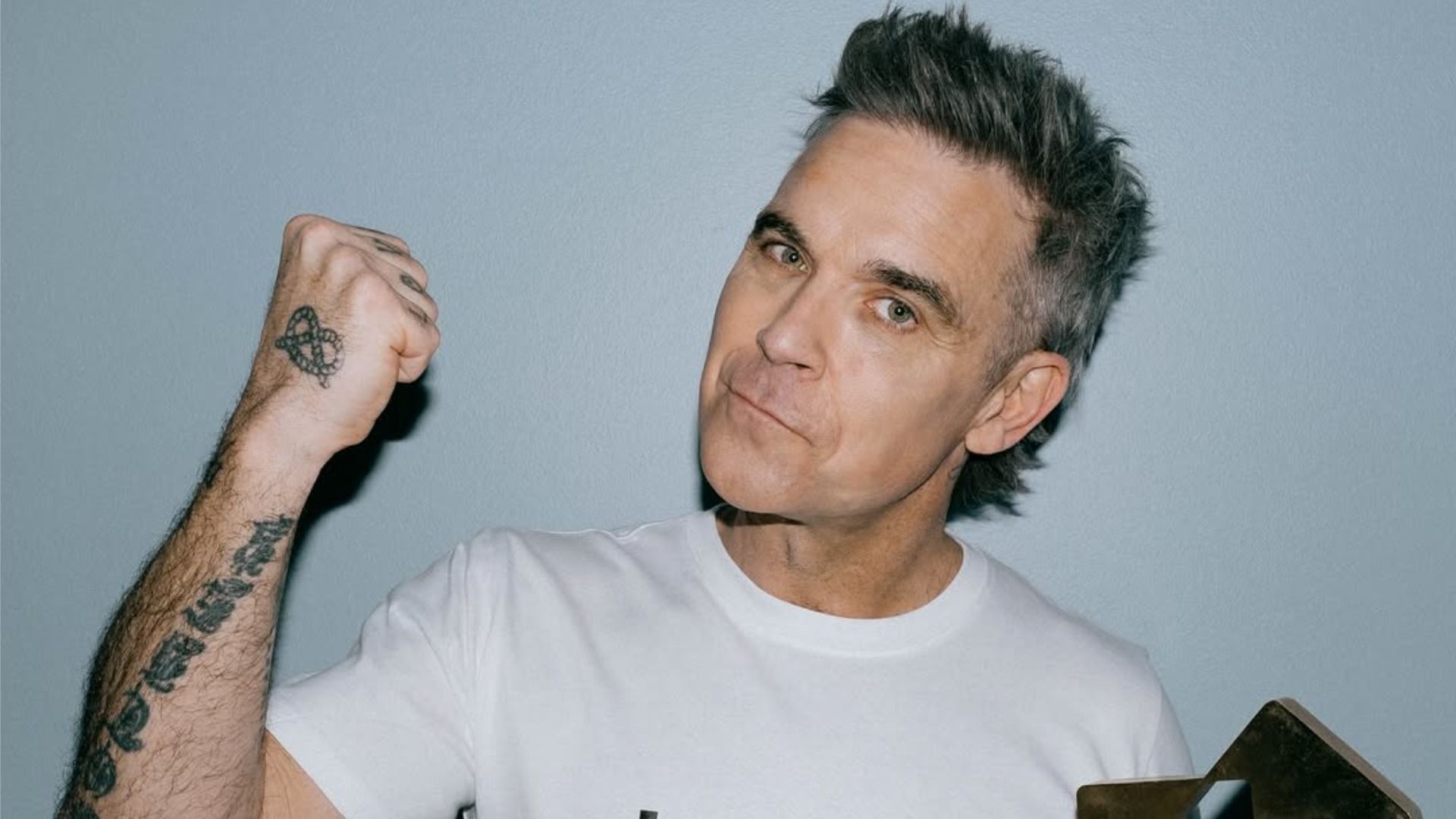 Cântărețul Robbie Williams și-a ridicat pumnul și zâmbește, uitându-se la cameră pe un fundal gri