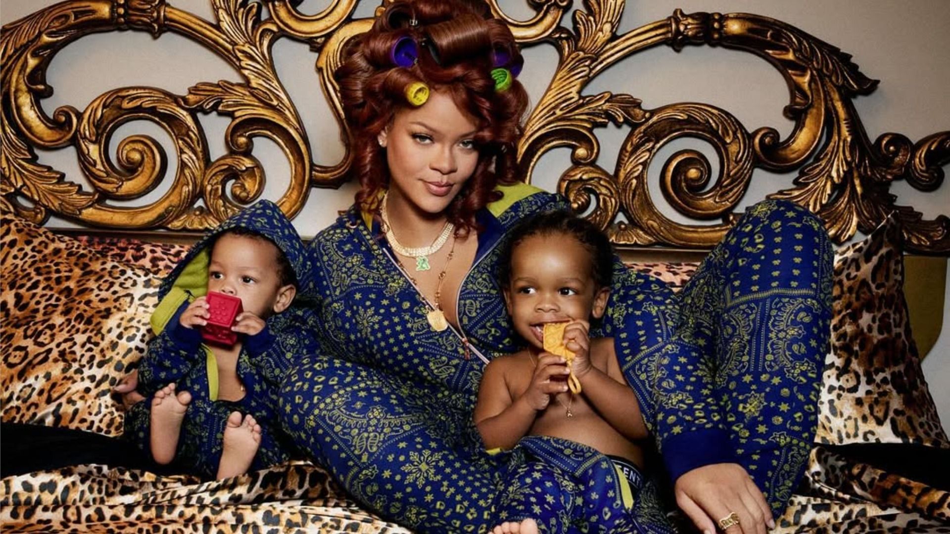 Cântăreața Rihanna este întinsă pe pat cu fiii ei RZA și Riot, privind la cameră cu un zâmbet