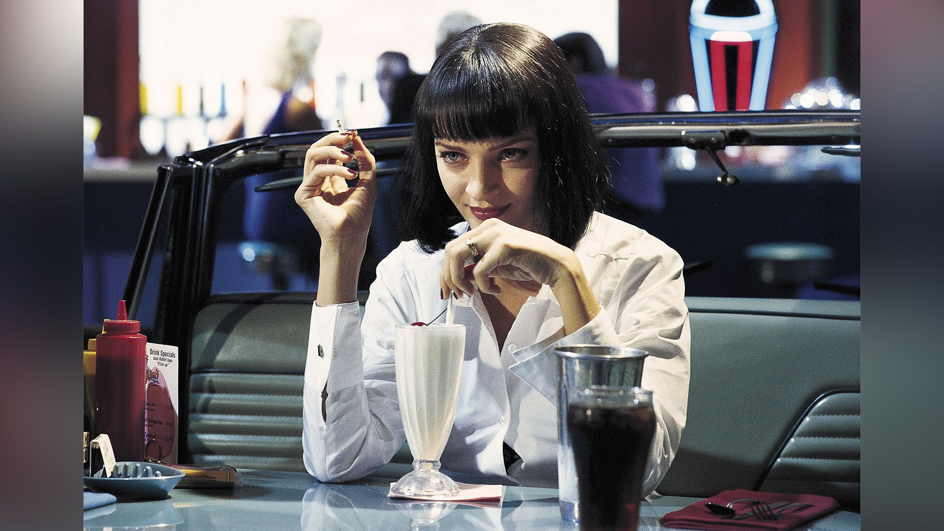 Uma Thurman într-o cămașă albă bea un milkshake într-o cafenea într-o scenă din filmul 'Pulp Fiction'