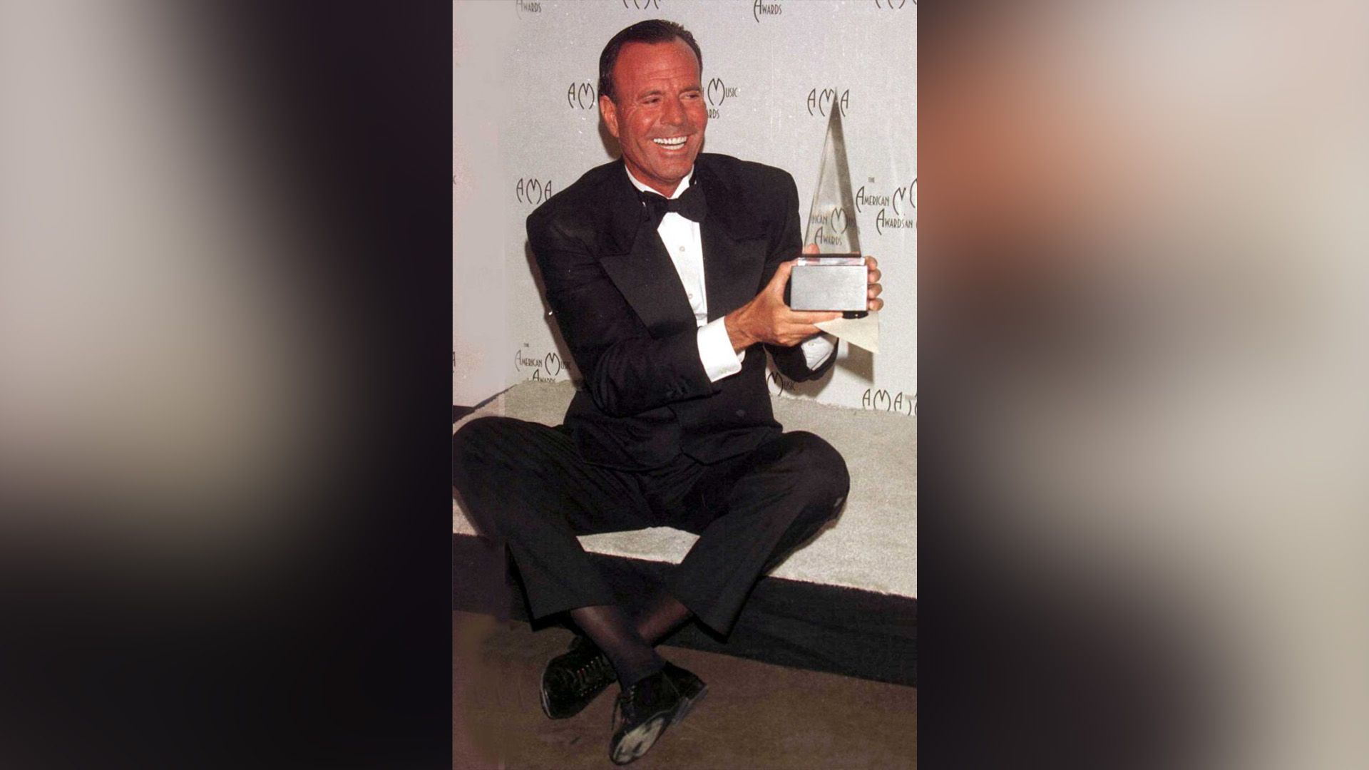 Julio Iglesias fericit, într-un costum negru, ținând în mâini trofeul American Music Awards