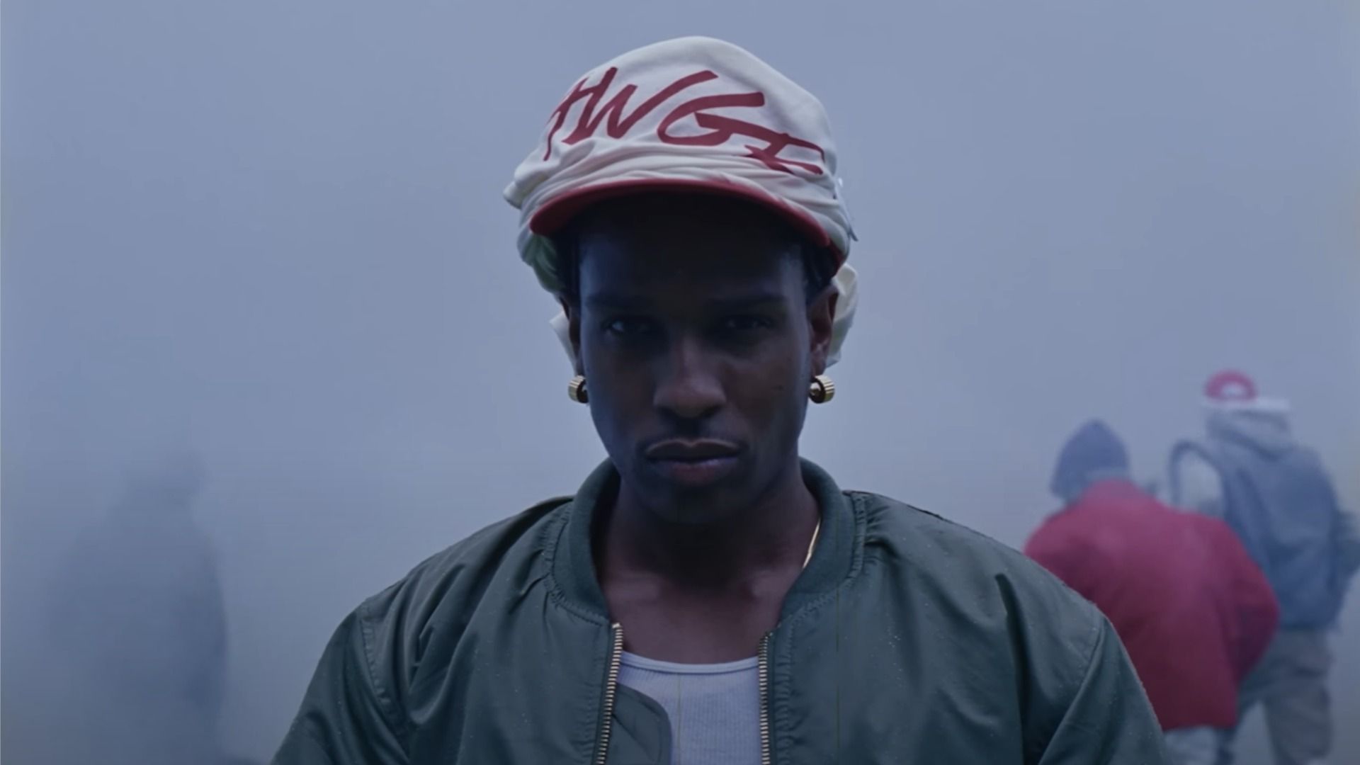Rapperul A$AP Rocky într-o jachetă bomber verde privește către cameră pe fundalul unei străzi fumurii