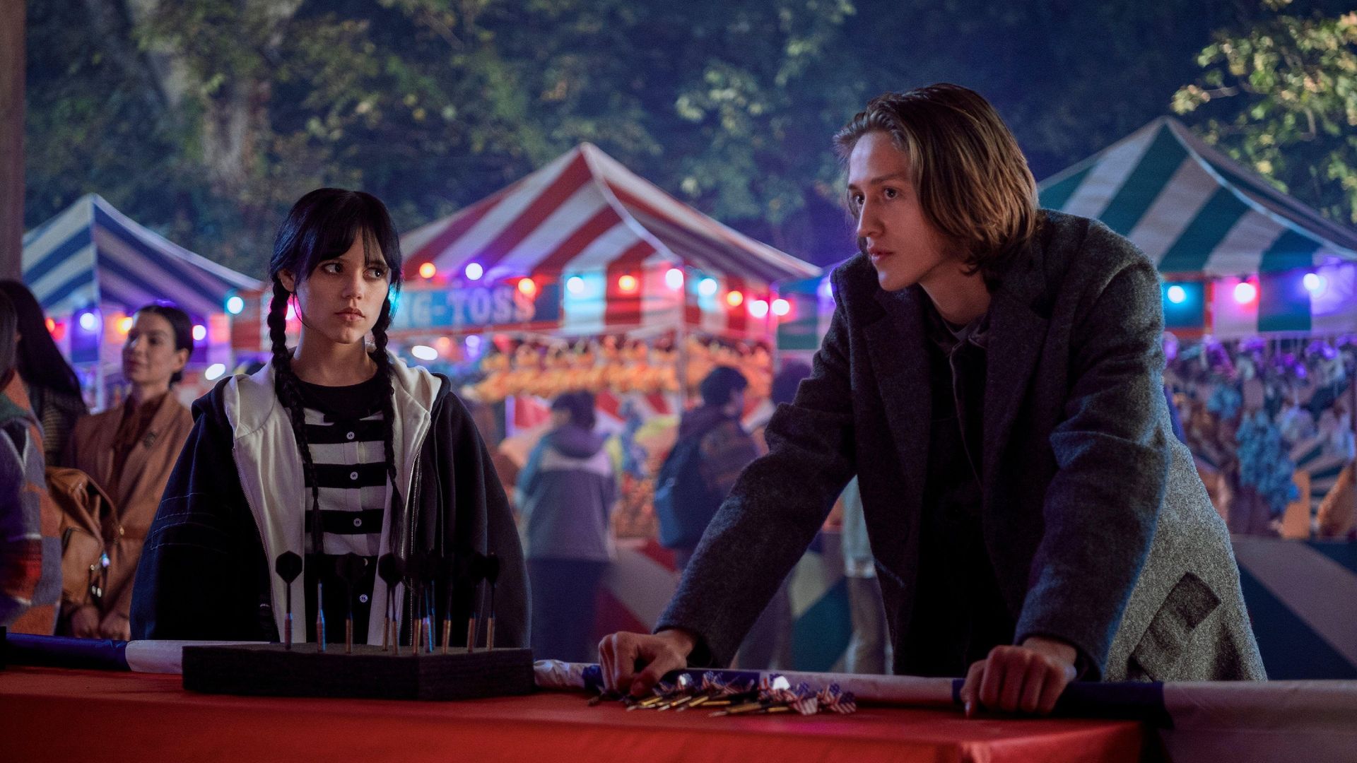 Jenna Ortega și Percy Hynes White în prim-plan cu un târg în fundal