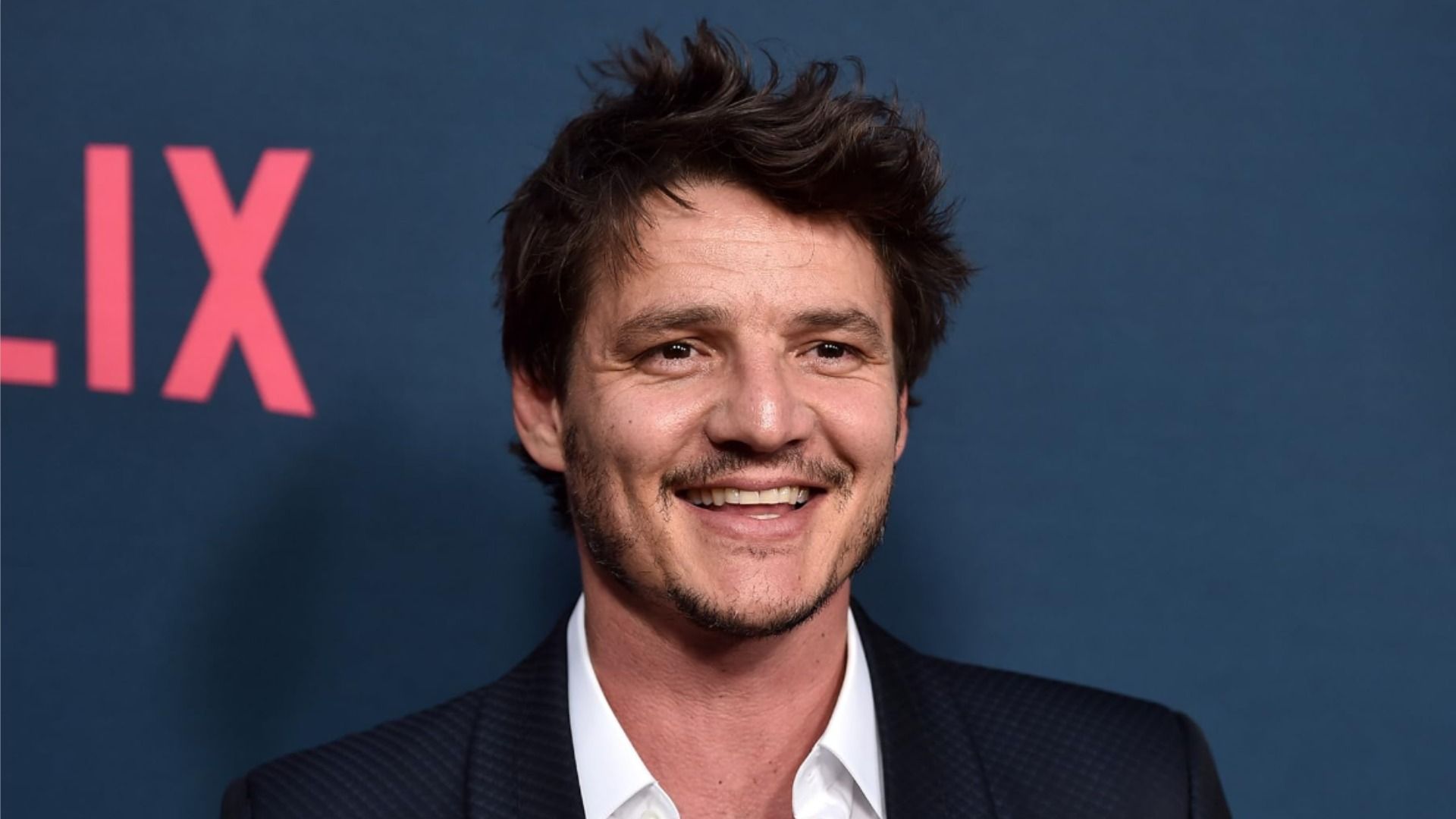 Actorul Pedro Pascal într-o jachetă neagră zâmbind pe un fundal albastru