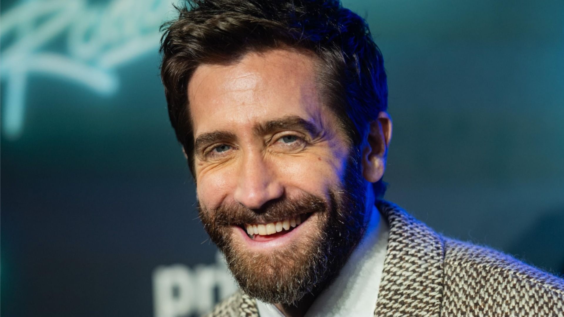 Actorul Jake Gyllenhaal într-un blazer gri zâmbește pe un fundal albastru