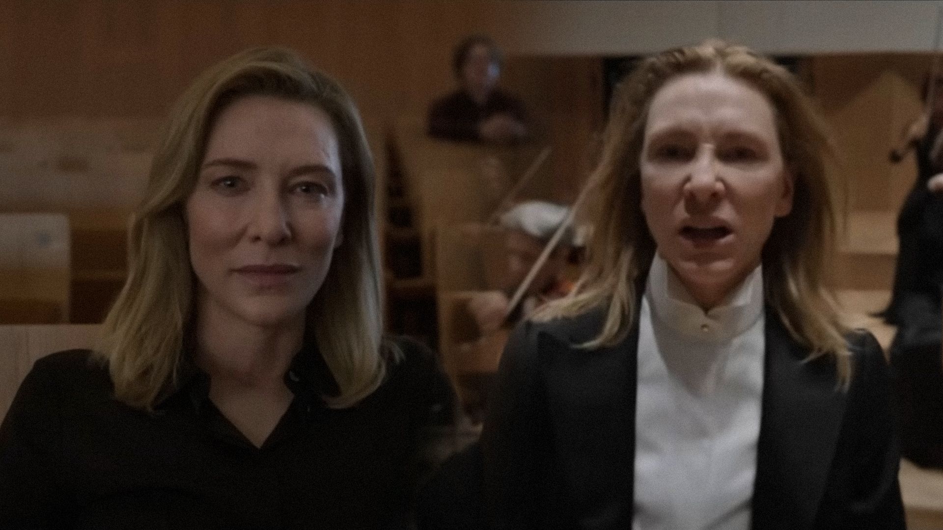 Actrita Cate Blanchett într-un costum negru mănuși amenințător la aparatul de fotografiat pe un fundal de violoniști