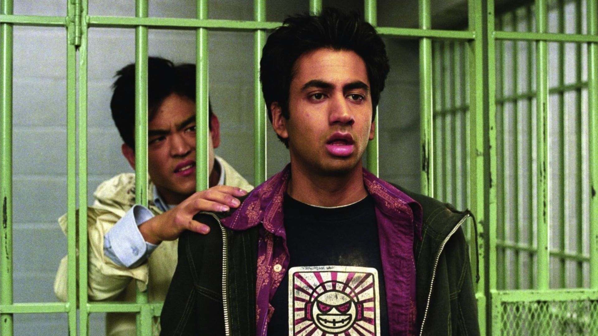 Actorul John Cho stă în spatele gratiilor verzi ale închisorii, actorul Kal Penn stă afară și se uită în lateral