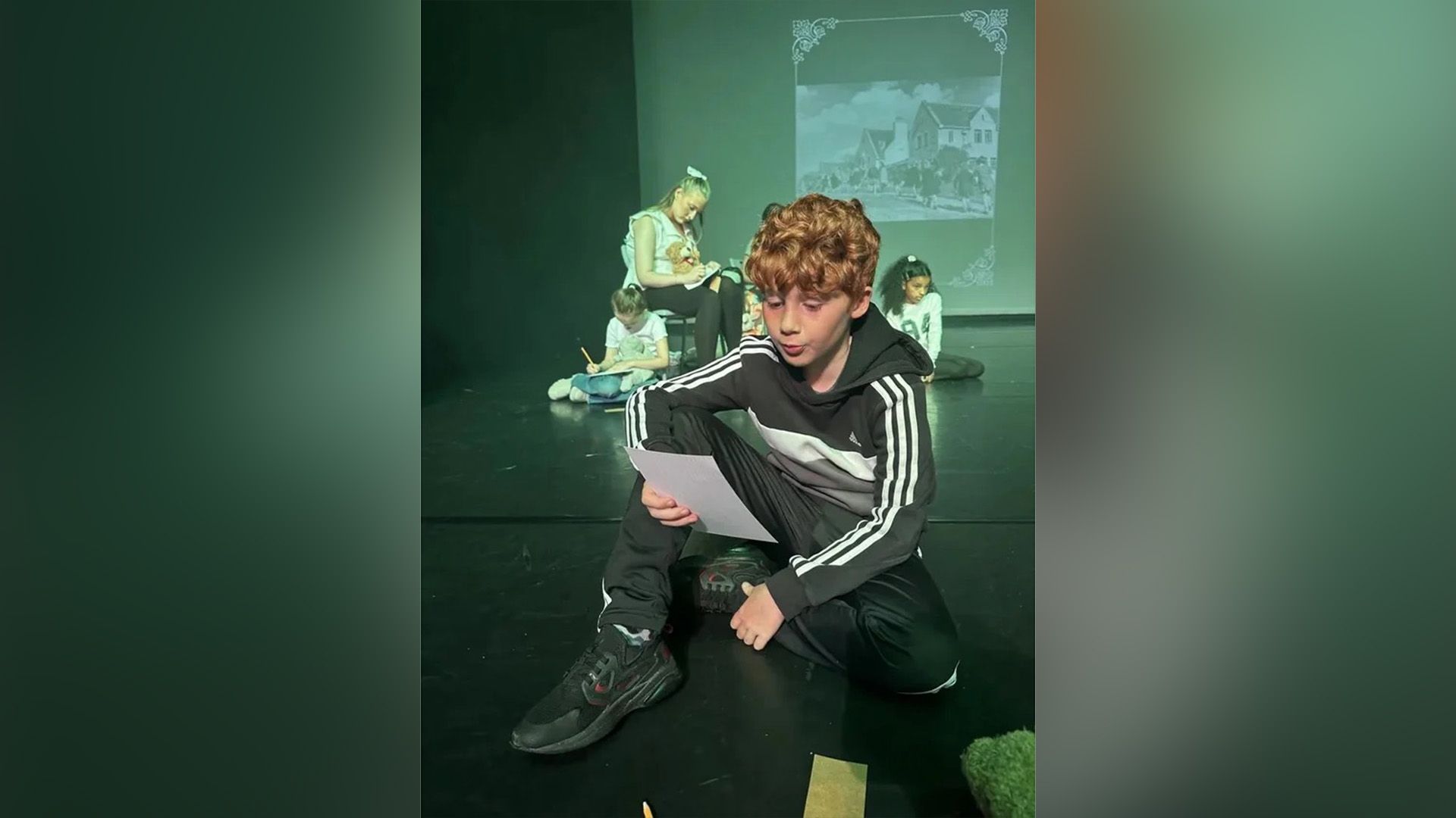 Alastair Stout într-un trening stând pe scena unui teatru și citind dintr-o foaie