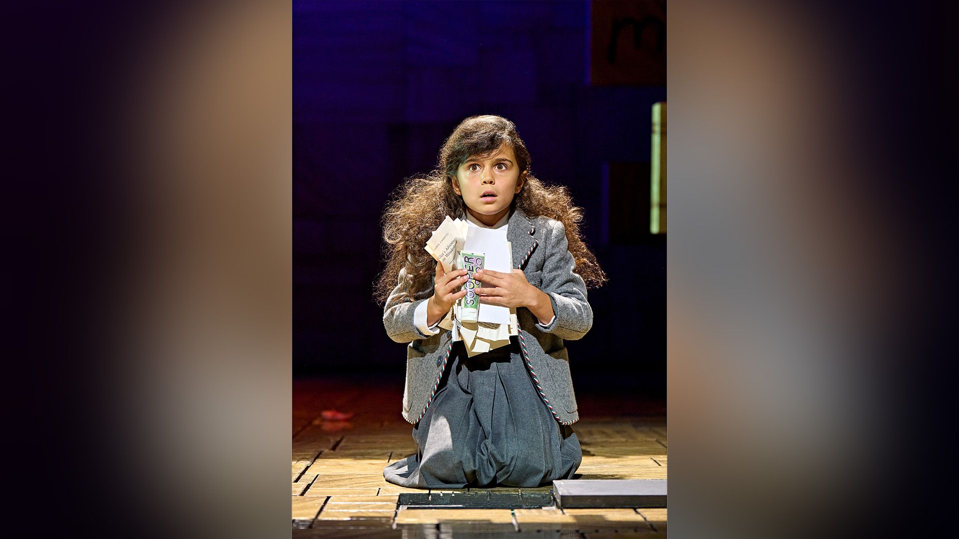 Arabella Stanton într-o rochie gri îngenunchind pe o scenă de teatru, ținând în mâini foi mototolite de hârtie