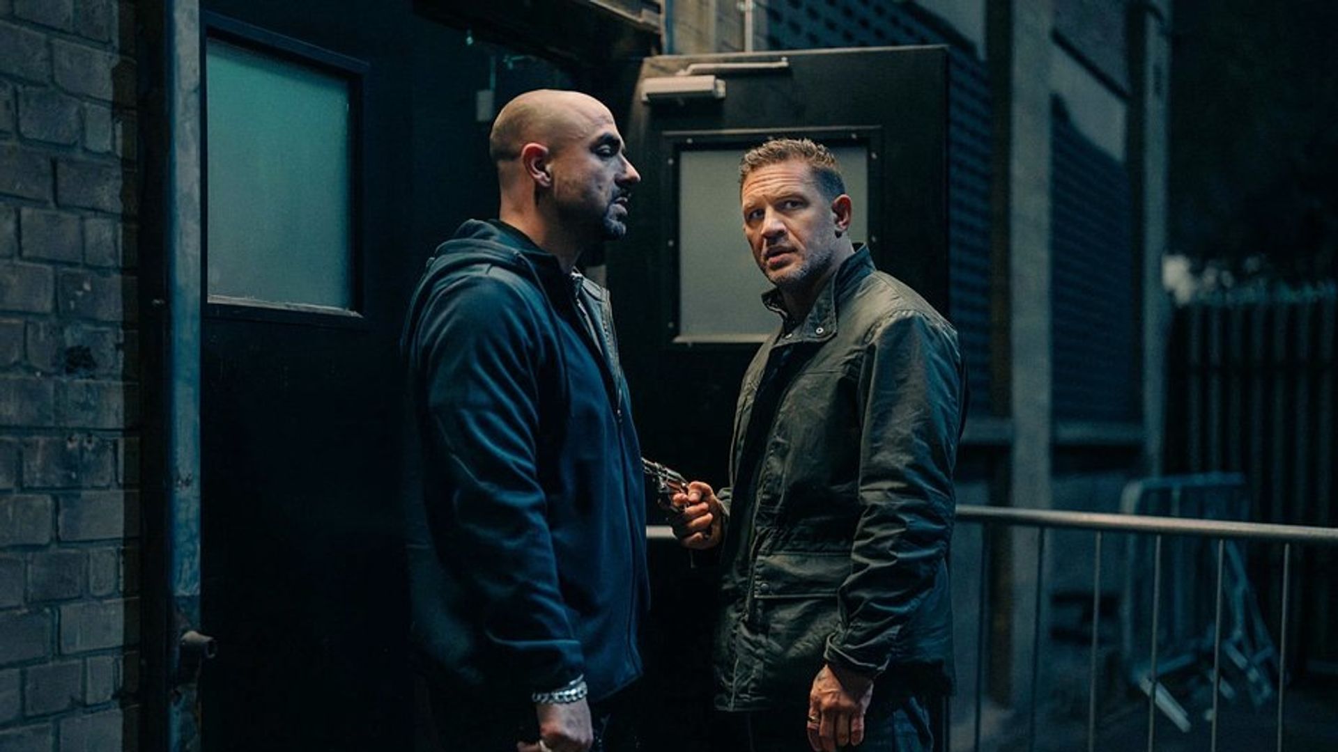 Tom Hardy și un alt bărbat în haine închise la culoare vorbesc lângă o ieșire neagră noaptea, unul dintre ei ține o armă