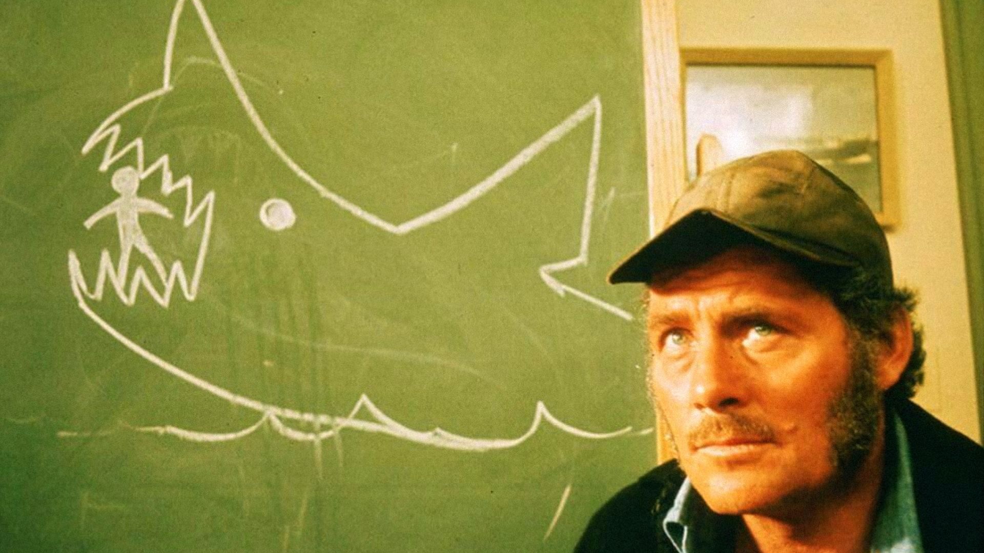 Actorul Robert Shaw cu o șapcă gri stă lângă o tablă cu un rechin desenat cu creta pe ea