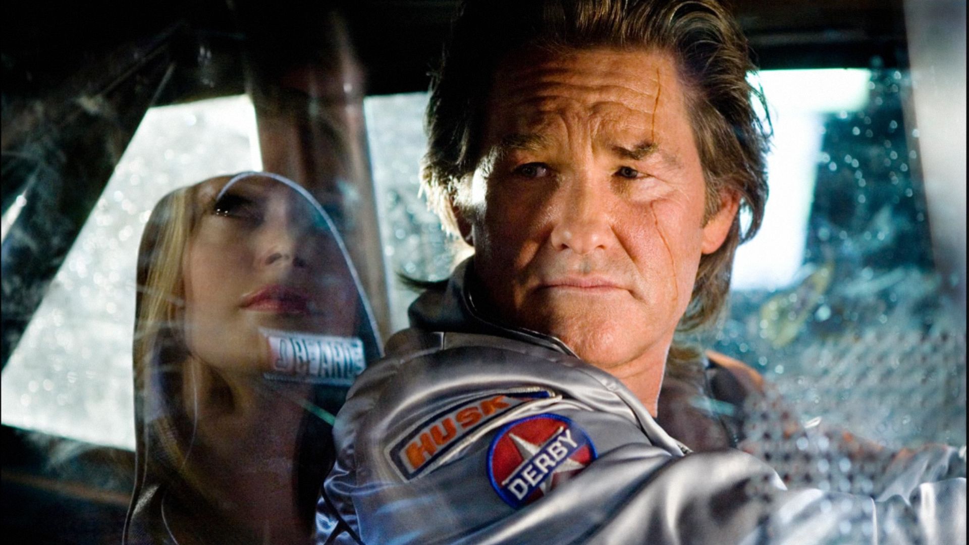 Actorul Kurt Russell într-o jachetă argintie stă la volanul unei mașini și privește într-o parte