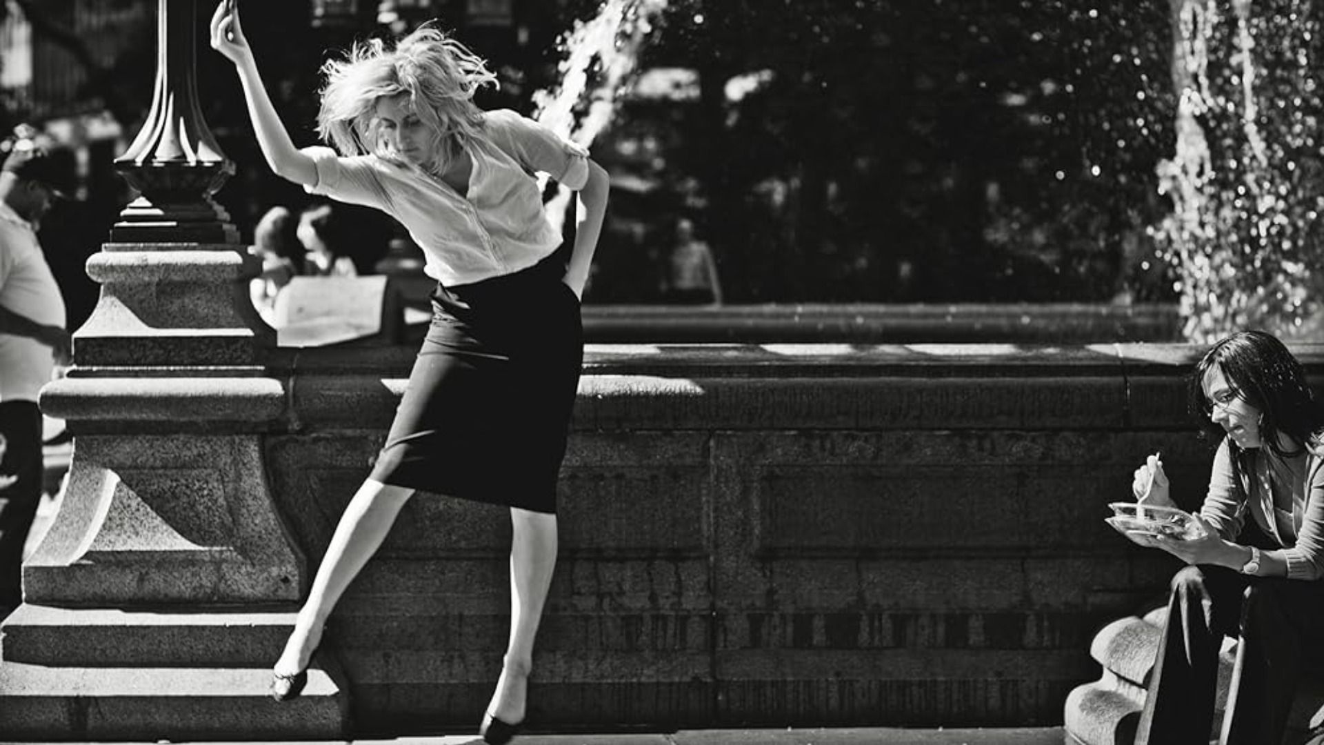 Actrița Greta Gerwig dansează într-o bluză albă și fustă neagră pe fundalul unei fântâni