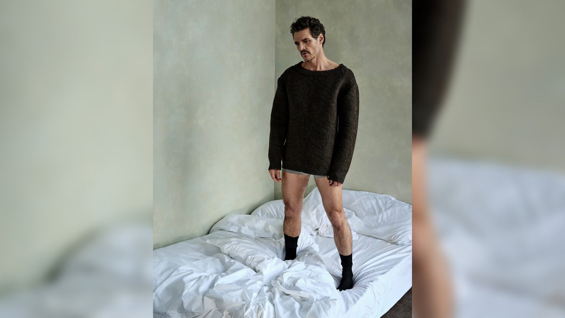 Pedro Pascal stând pe pat în pulover și lenjerie intimă