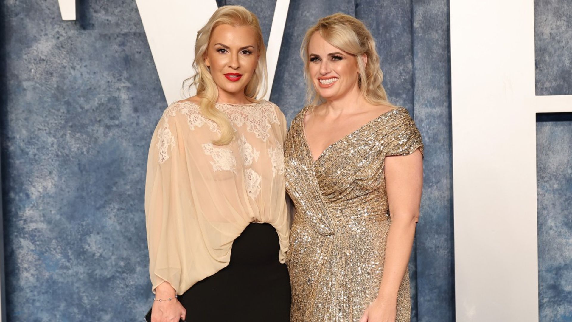 Rebel Wilson și Ramona Agruma stând în rochii pe un fundal deschis