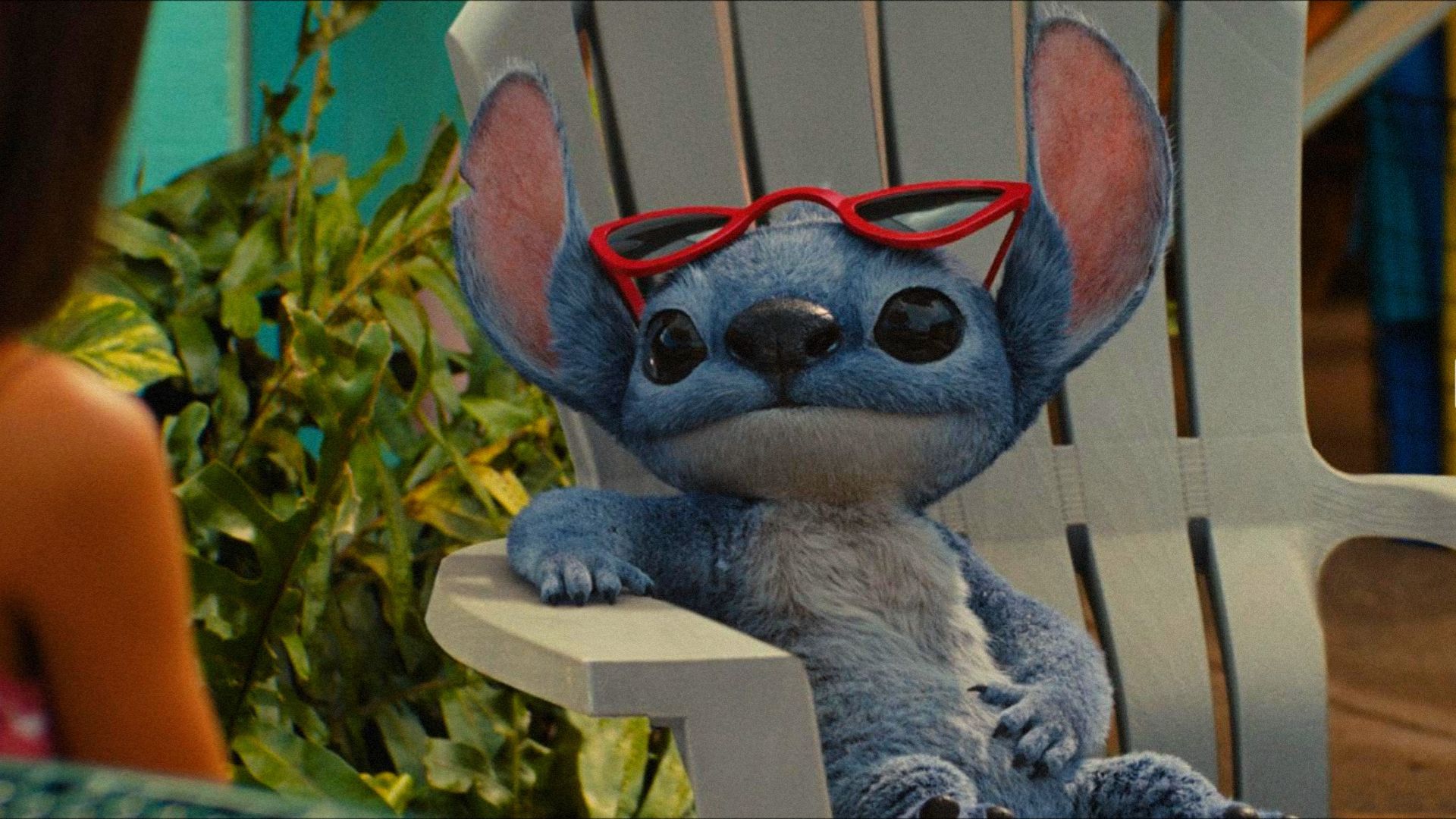 Alienul albastru Stitch stă pe un scaun de plajă purtând ochelari și se uită într-o parte