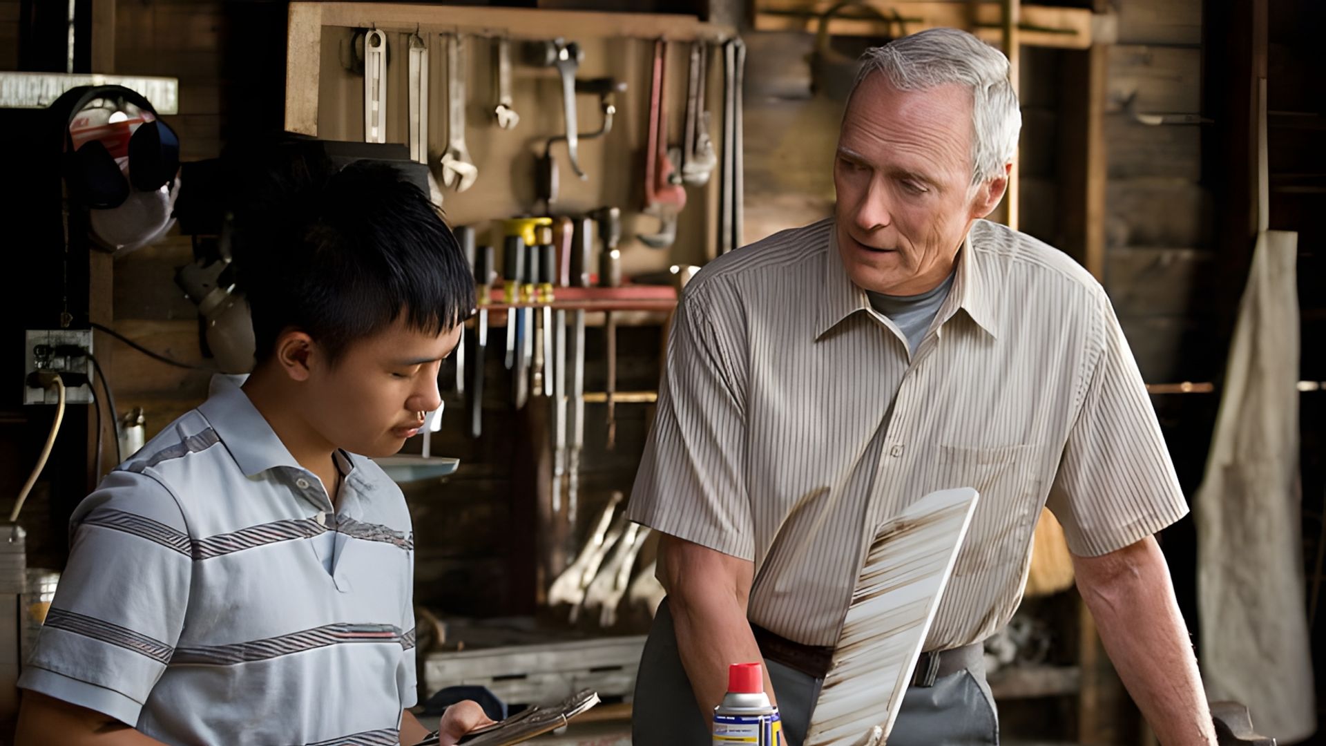 Clint Eastwood și un adolescent lucrează într-un atelier, discutând despre o unealtă pe fundalul unui banc de lucru cu unelte atârnate