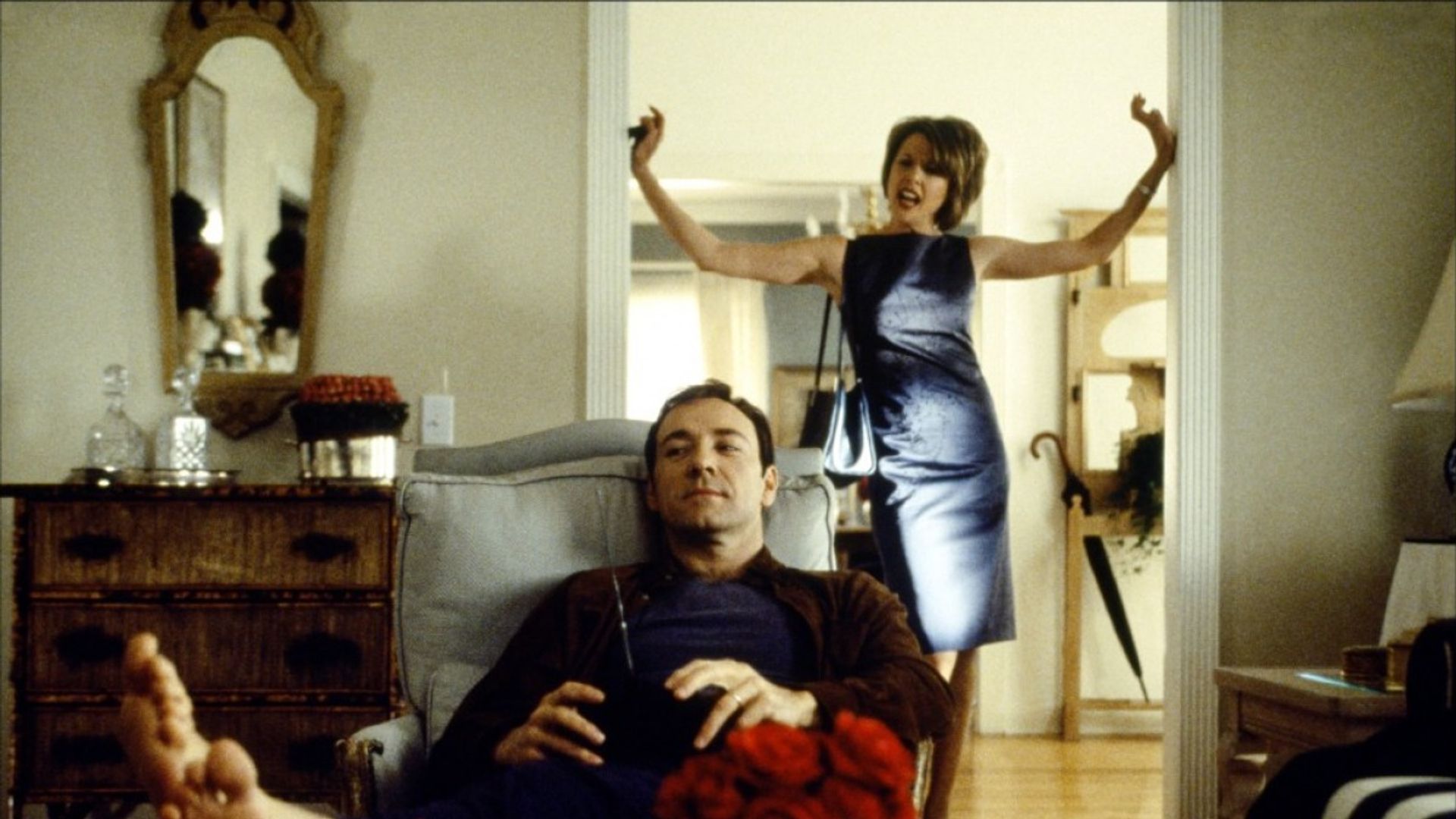 Kevin Spacey stă relaxat într-un fotoliu cu telecomanda în mână în timp ce o femeie în rochie de seară din spatele lui gesticulează emoțional în pragul ușii