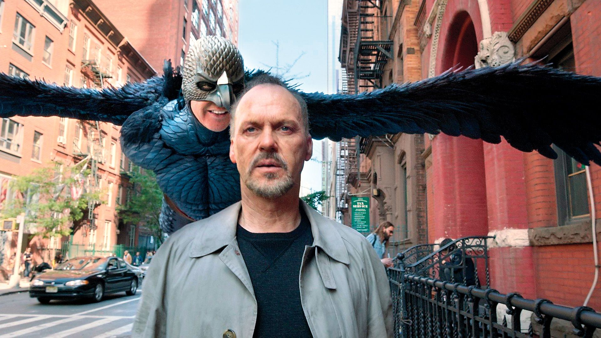 Michael Keaton merge pe stradă în timp ce în spatele lui plutește un personaj fantastic cu aripi și mască de pasăre