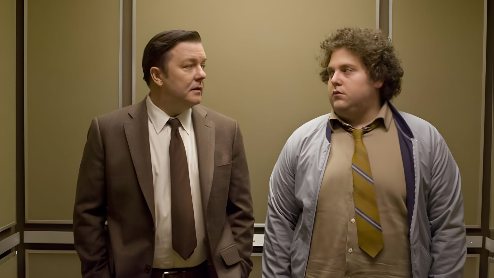 Ricky Gervais și Jonah Hill în costume de afaceri stând într-un lift, privind unul la celălalt cu expresii serioase pe față
