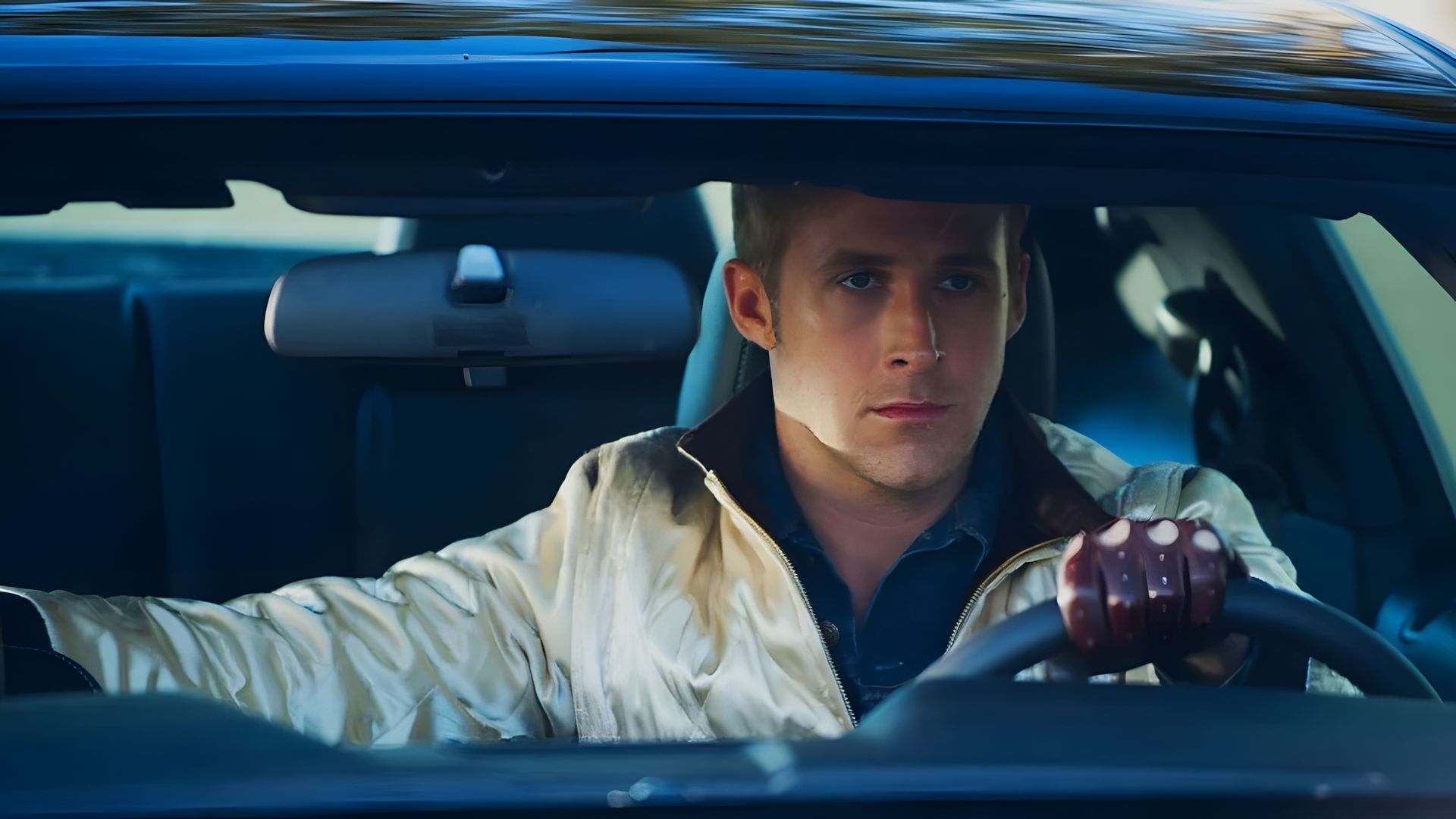 Ryan Gosling într-o jachetă de satin și mănuși de piele stă la volanul unei mașini, privind înainte cu o expresie concentrată