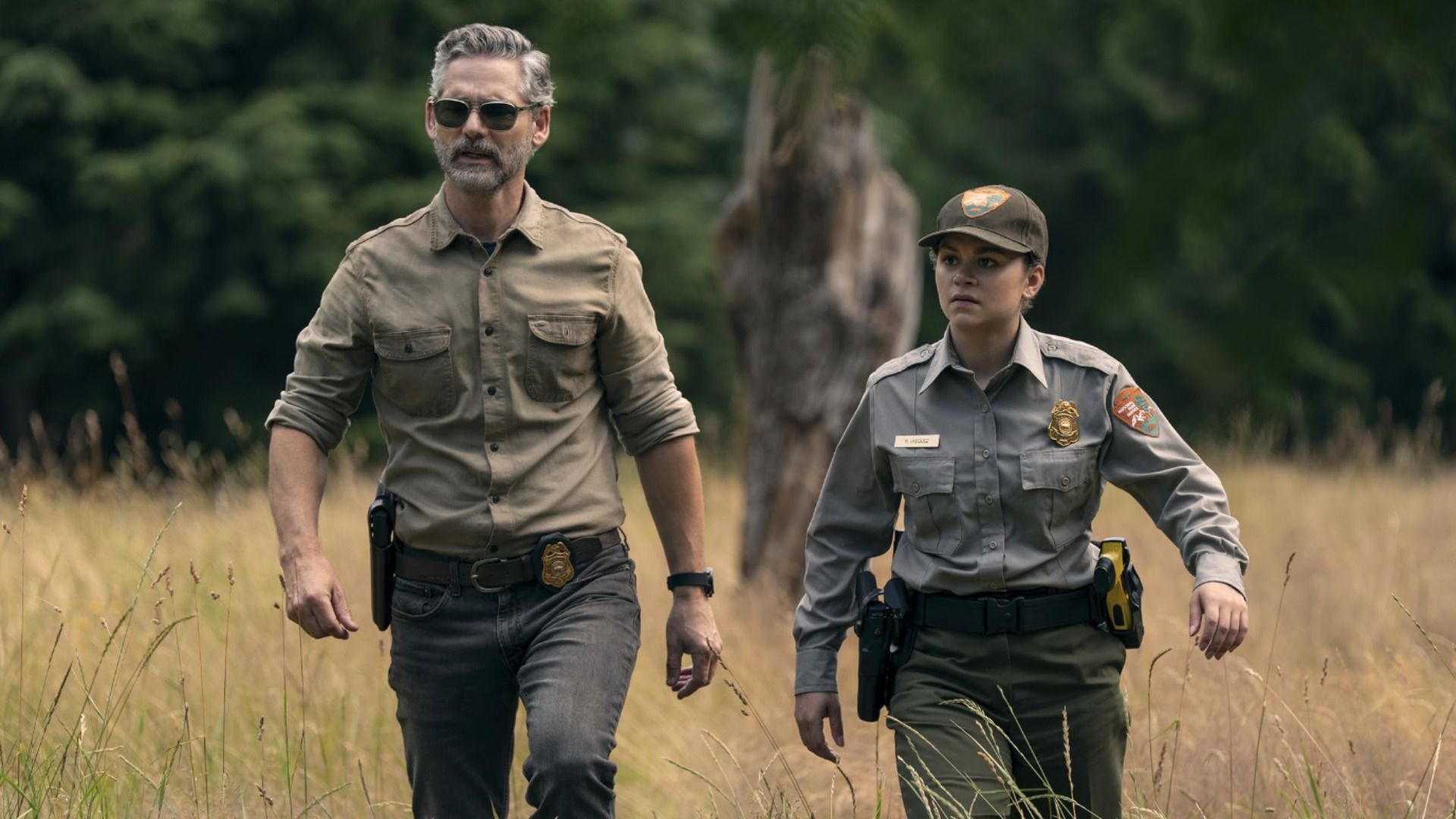 Actorul Eric Bana într-o cămașă verde se plimbă cu o polițistă peste iarba galbenă cu copaci în fundal